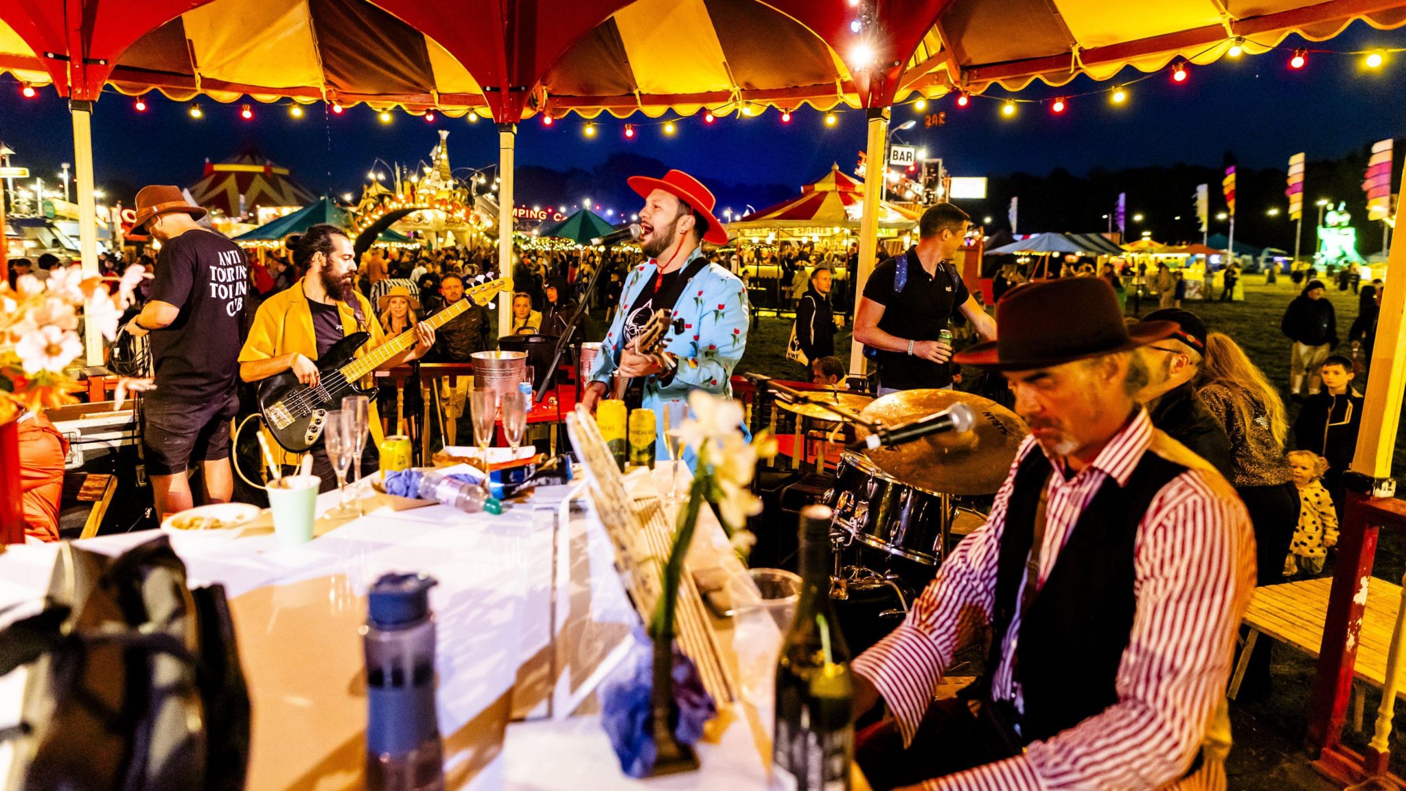 A man in a red hat and blue jacket stands, singing into a microphone. He is holding a guitar. Next to him is another man in a yellow jacket, who also has a guitar. To the right, there is a man in a red hat and red and white striped shirt, sitting at a piano. The sky is dark in the background and there are hundreds of people walking on a field