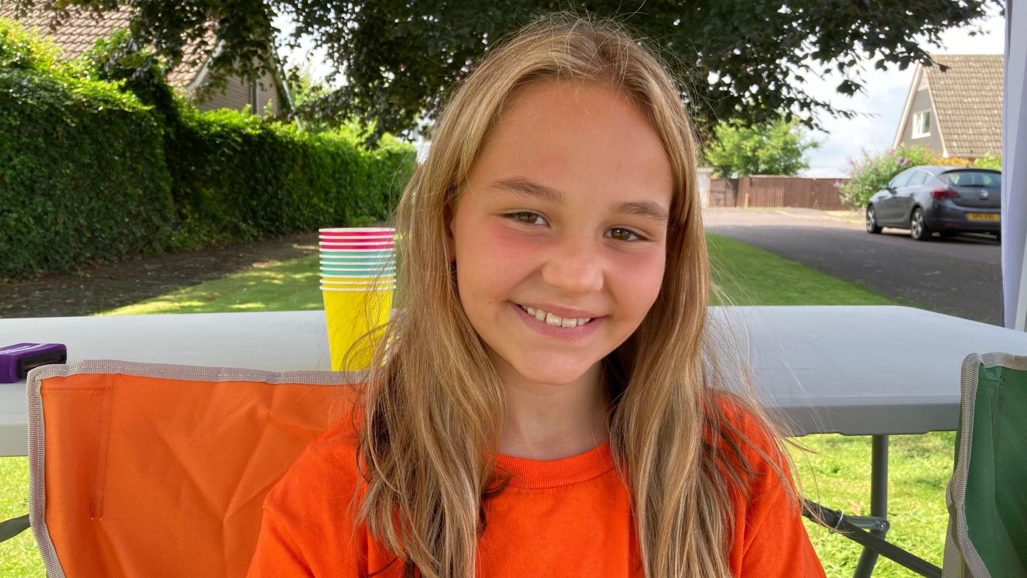 Shyla Brown smiles at the camera while sat outdoors on an orange camping chair. She has brown eyes, blonde hair and is wearing a bright orange t-shirt. 