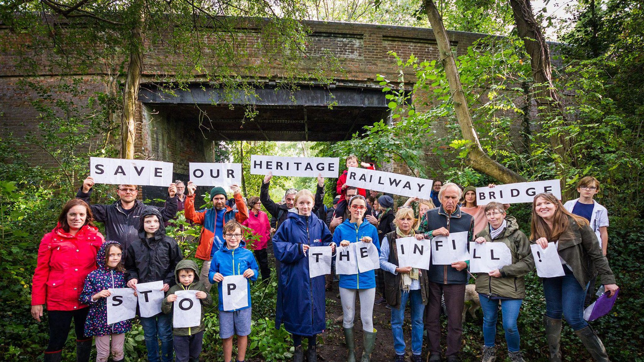 A group of campaigners of all ages stand in front of the bridge in Barcombe holding bits of paper which read "Save our heritage railway bridge" and "stop the infill"