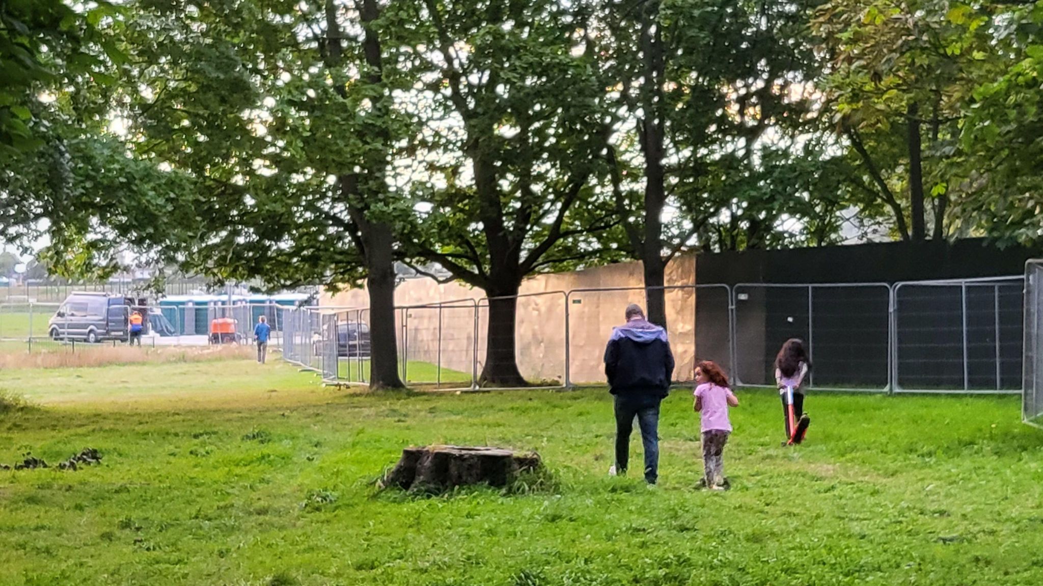 Two children and man walk through the park near temporary metal fences in August 2023