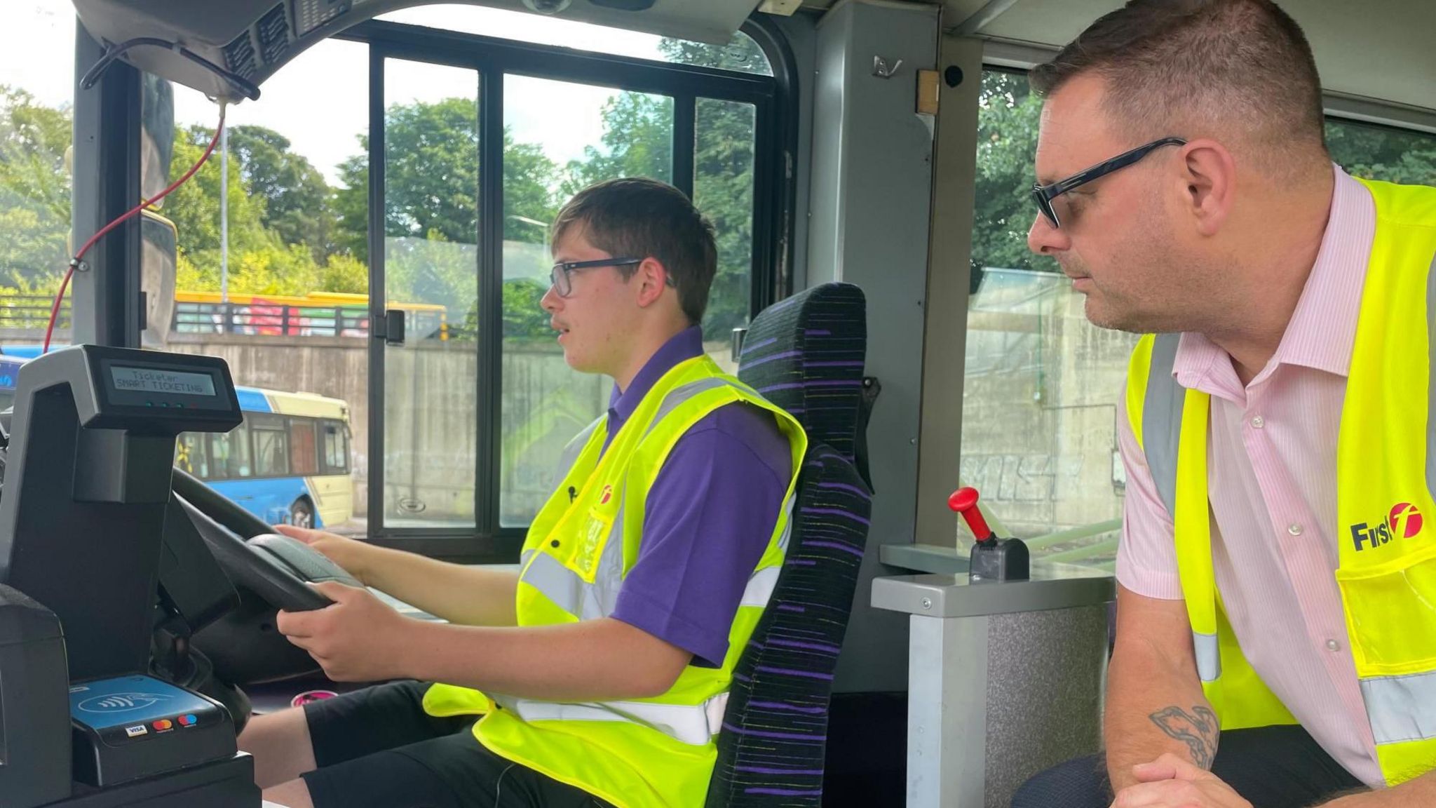 A young man sits in the driving seat of a bus whilst an older man sits next to him watching closely