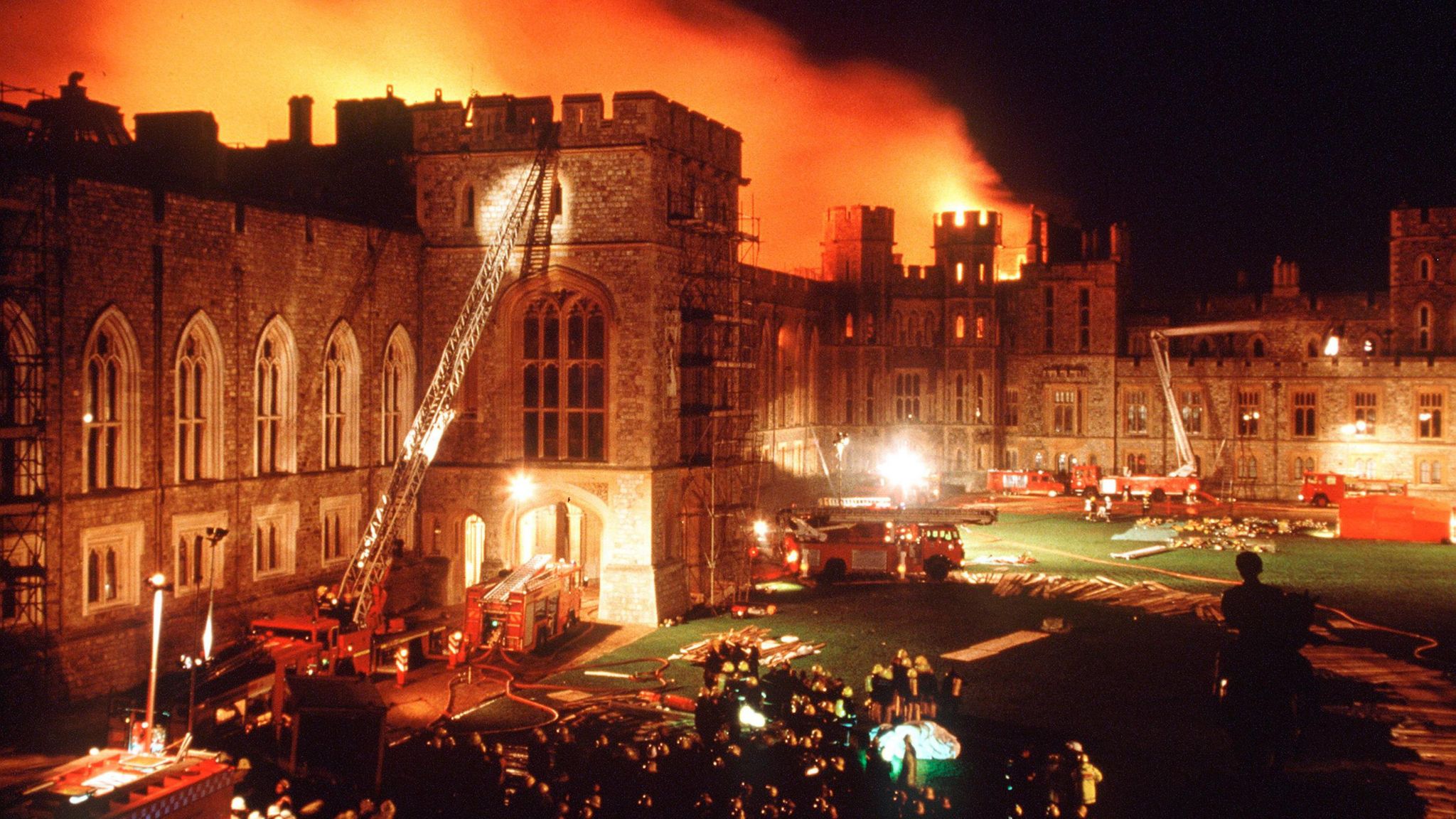 A large castle on fire in the distance, with large crowds of firefighter and fire engines in front tackling the blaze