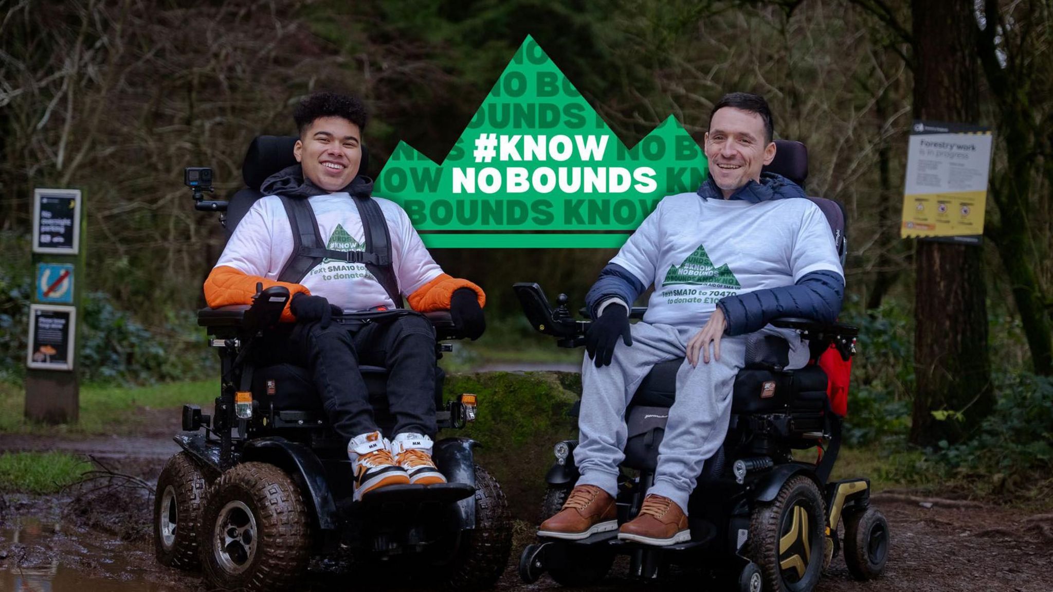 Maxwell McKnight and Josh Wintersgill side by side in their wheelchairs in a woodland setting with a sign carrying the name of their challenge, Know No Bounds