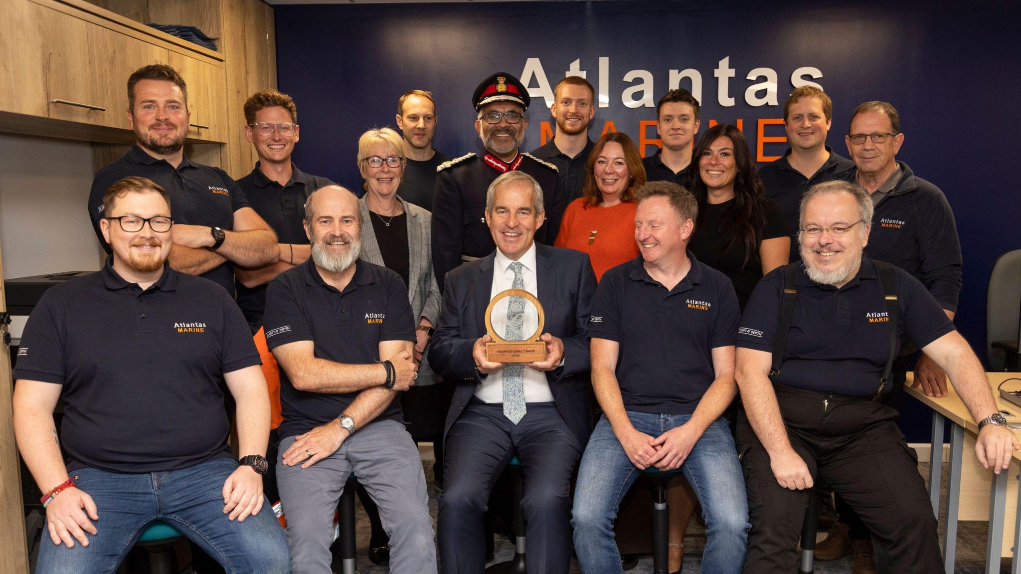 Charlie Foll from Atlantas Marine sits alongside team members whilst holding King's Award presented to him by Lord-Lieutenant of Somerset who is stood behind him.