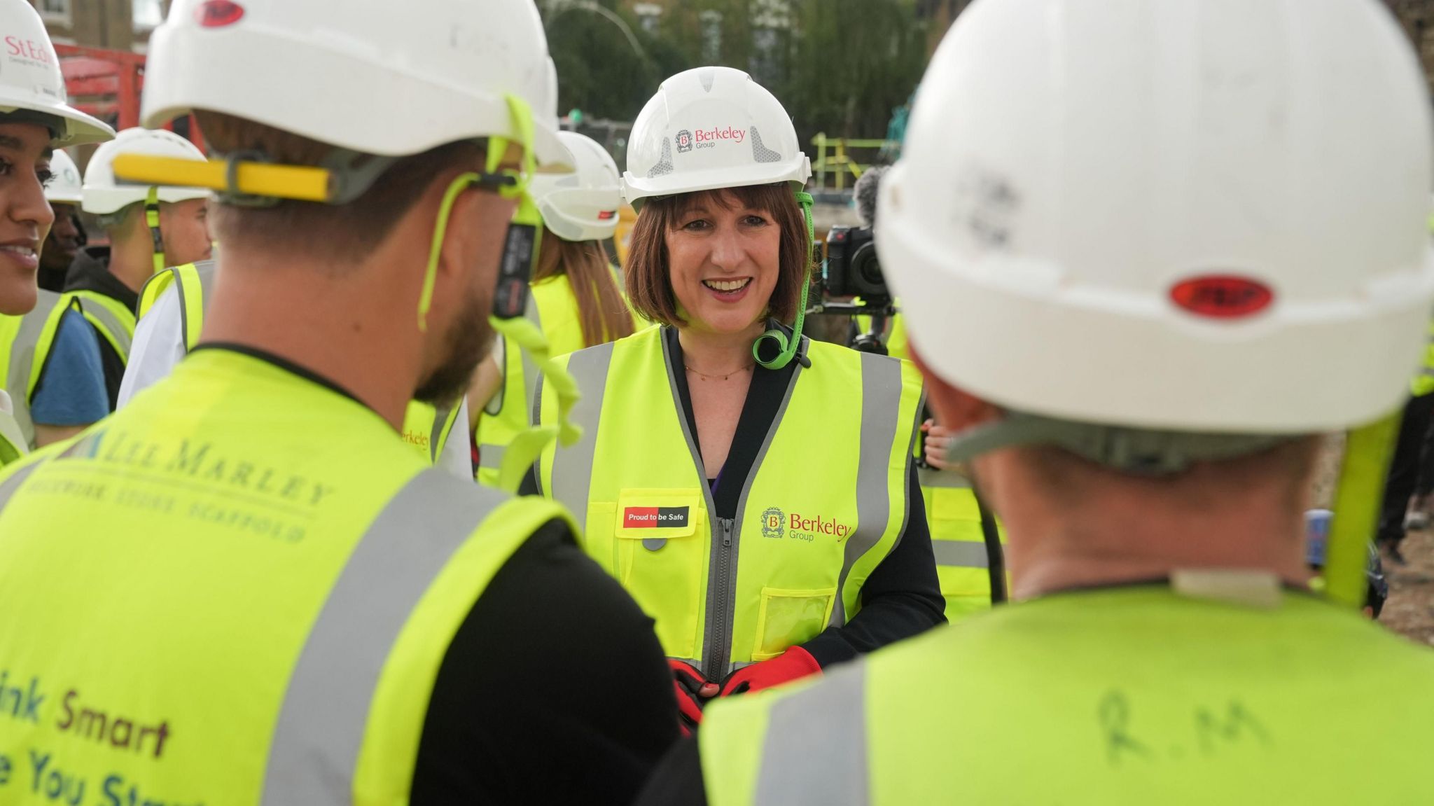 A smiling Rachel Reeves wearing a high-visibility jacket and white hard hat speaks to two construction workers at a site