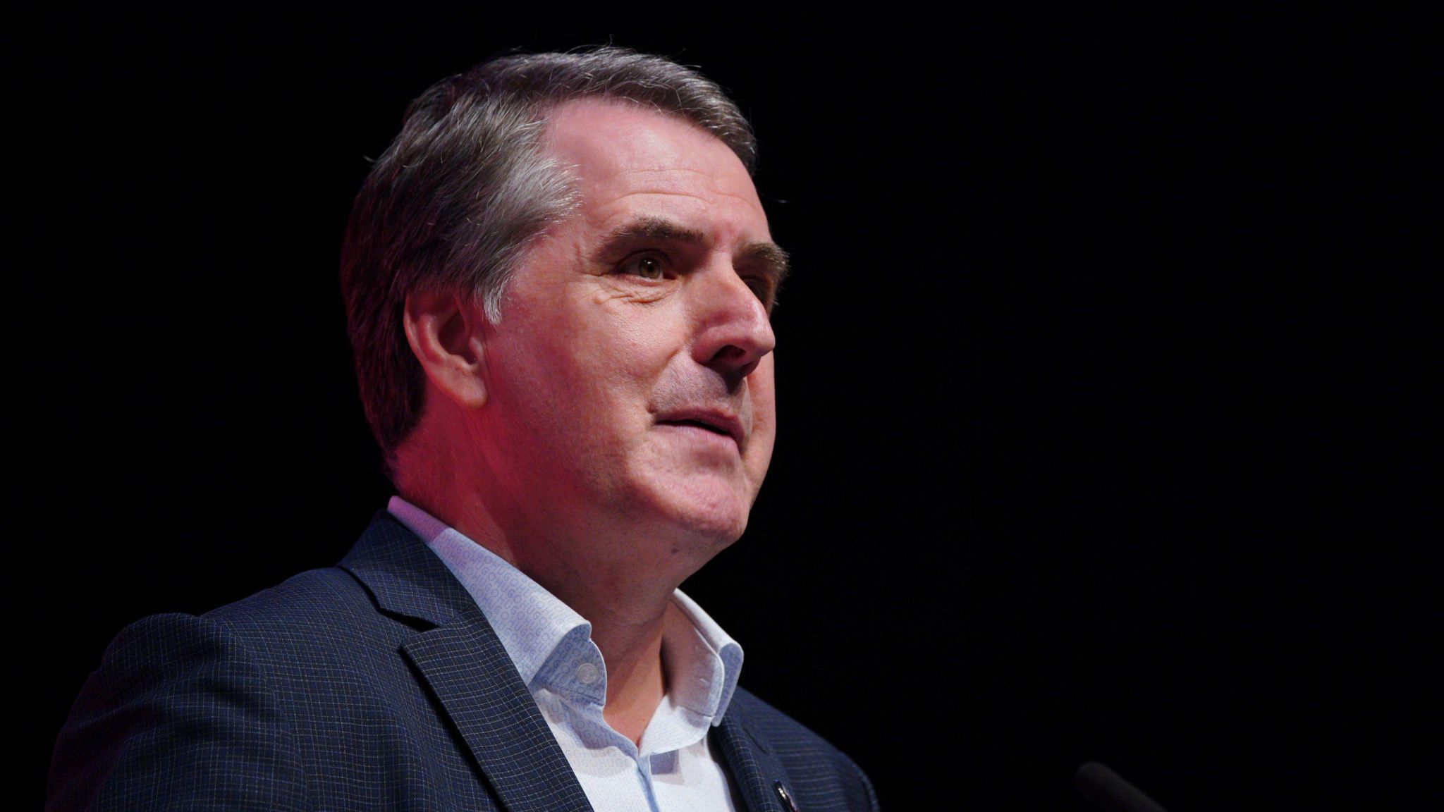 Mayor Steve Rotheram standing next to a dark wall. He has short dark grey hair. He is wearing a navy suit with a pale blue shirt. 