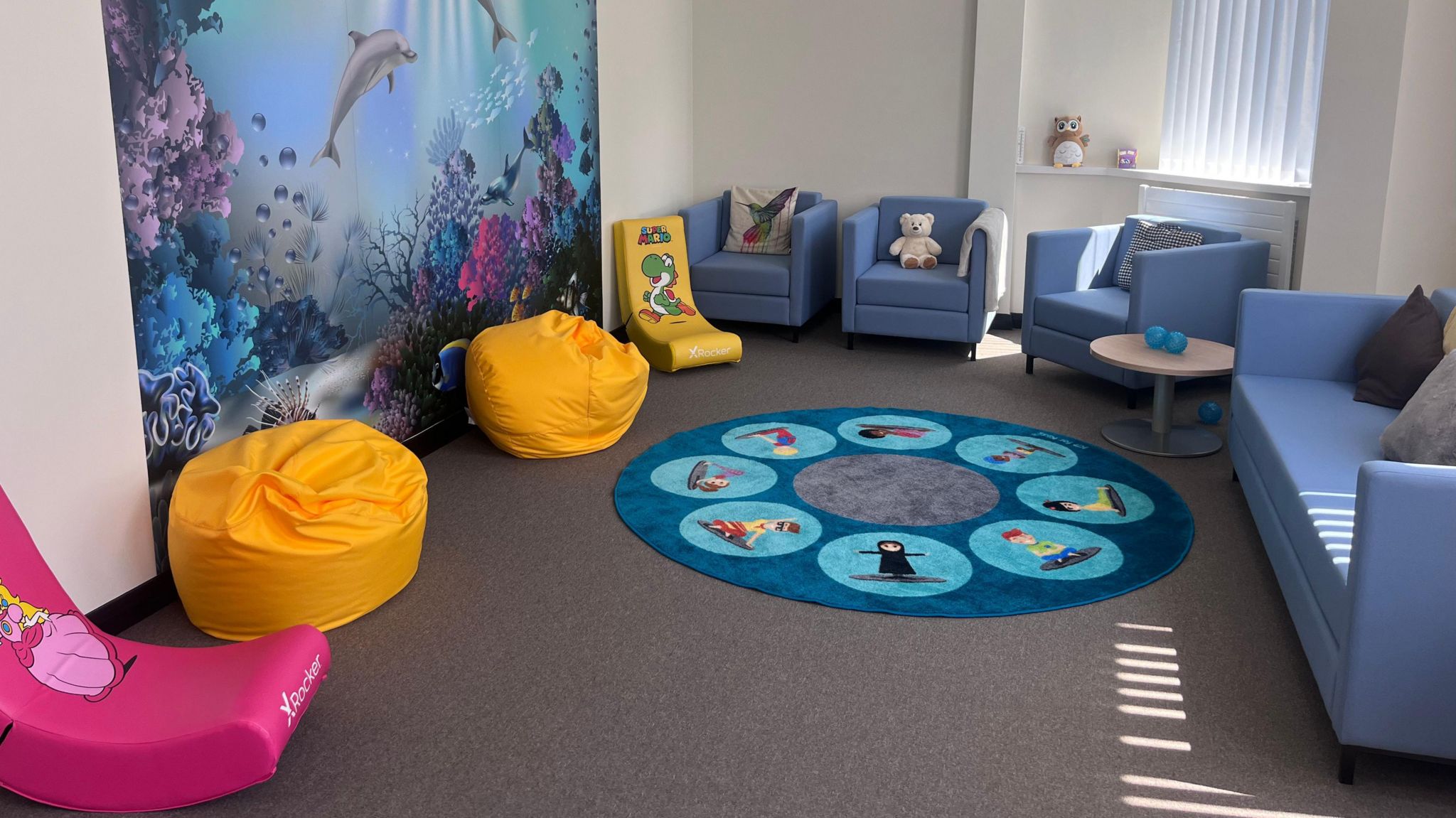 The children's room at the facility - there's a sealife picture on the wall, bright yellow bean bags and pink and yellow rocker chairs. There's also a rug and a teddy bear on one of the blue chairs