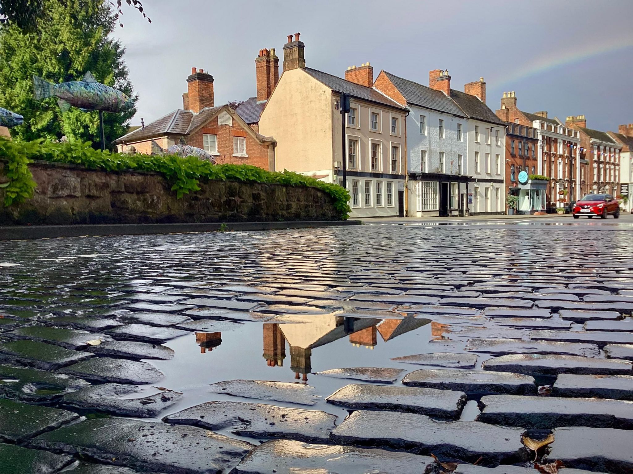 A ground-level shot focusing on brick cobbles with a puddle reflecting the three-storey, 18th Century buildings behind.