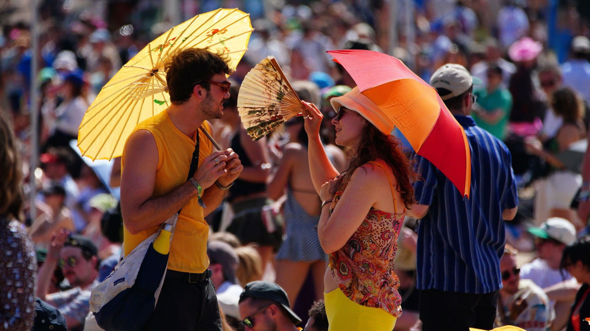Image of a man and woman at Glastonbury Festival. They are holding sun parasols, wearing sunglasses and having a conversation. The woman is holding a fan.  