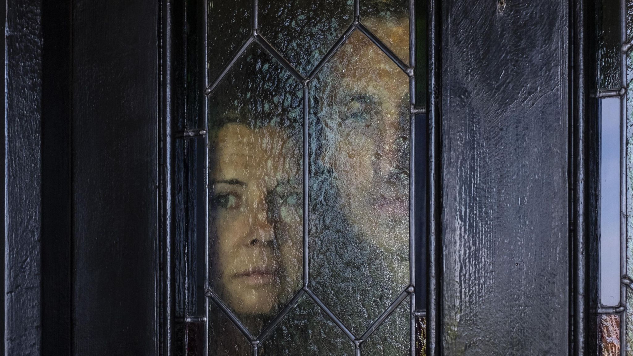 Two faces look through a pane of glass with hexagonal panels on a black front door. On the right of the pane, there is a woman looking to the side. Next to her is half the face of a man - this is more out of focus. 