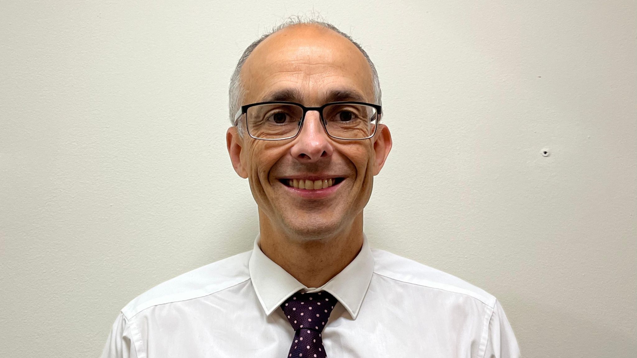 Chief Constable, Paul Sanford smiling. He is wearing large framed, rectangular glasses and has little hair. He is dressed in a white shirt and a dark purple tie with gold spots on it