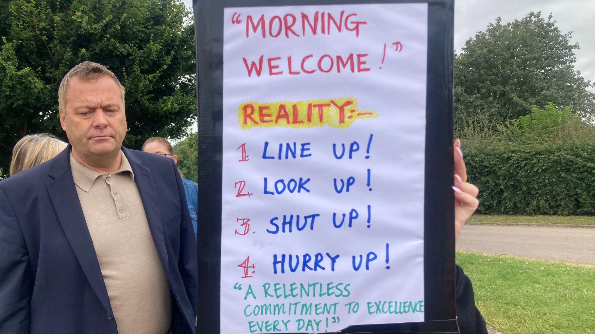 A picket line member holding a placard which reads: "Morning Welcome! Reality:- 1. Line up! 2. Look up! 3. Shut up! 4. Hurry up! 'A relentless commitment to excellence every day!'"