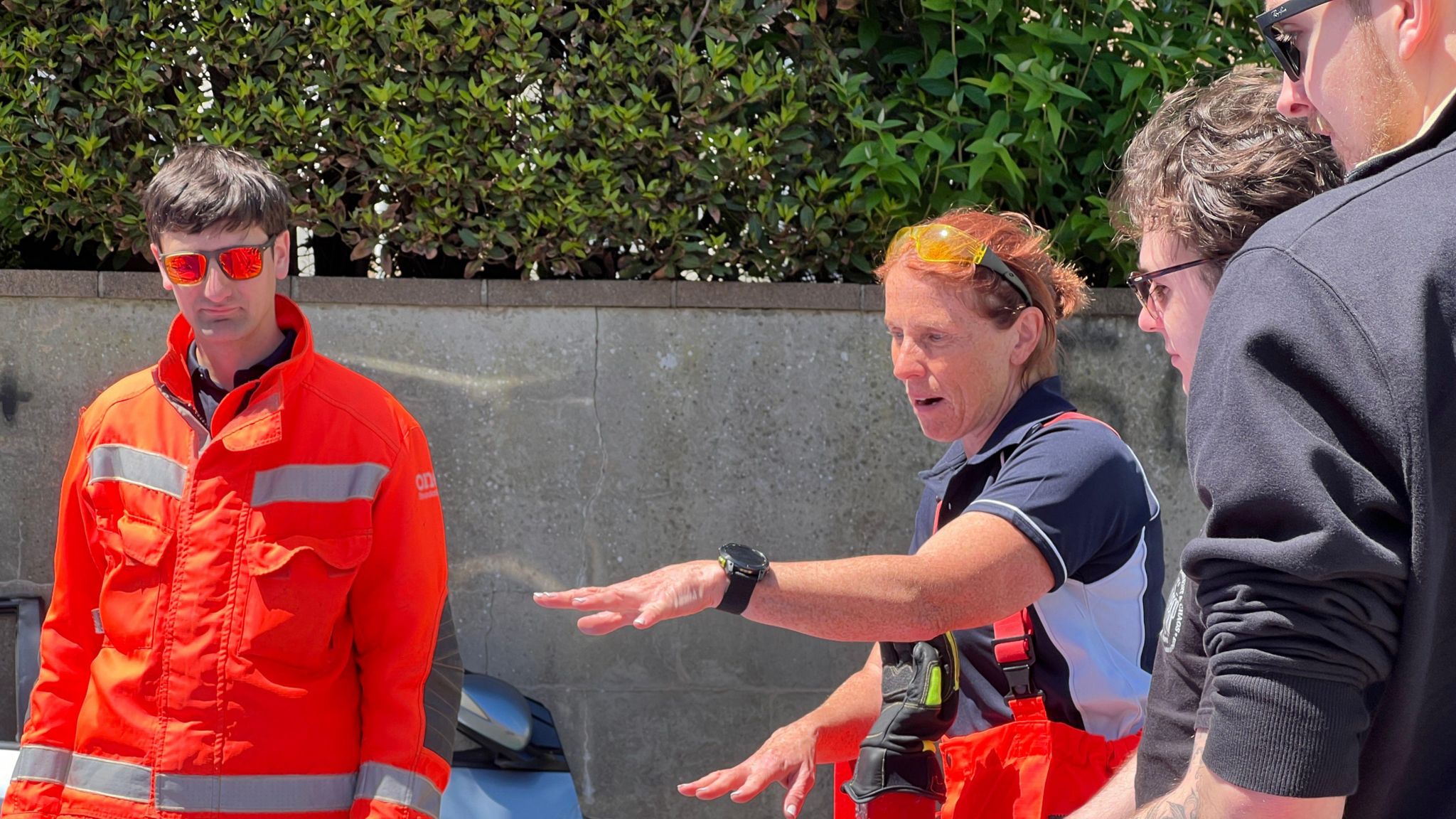 Helen is gesturing to the ground as two men stand next to her watching what she says and another member of Normandy Rescue is to her left in full high vis overalls