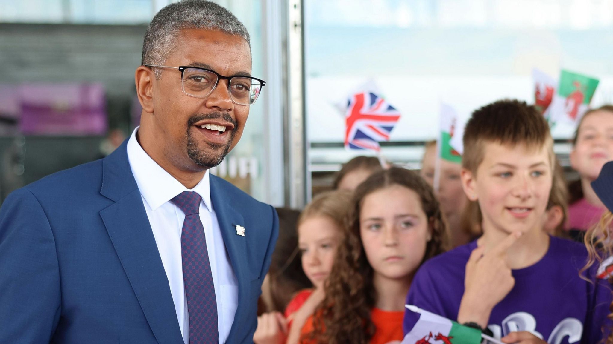 Vaughan Gething at the Senedd standing alongside children waving Wales and Union Jack flags at the Senedd