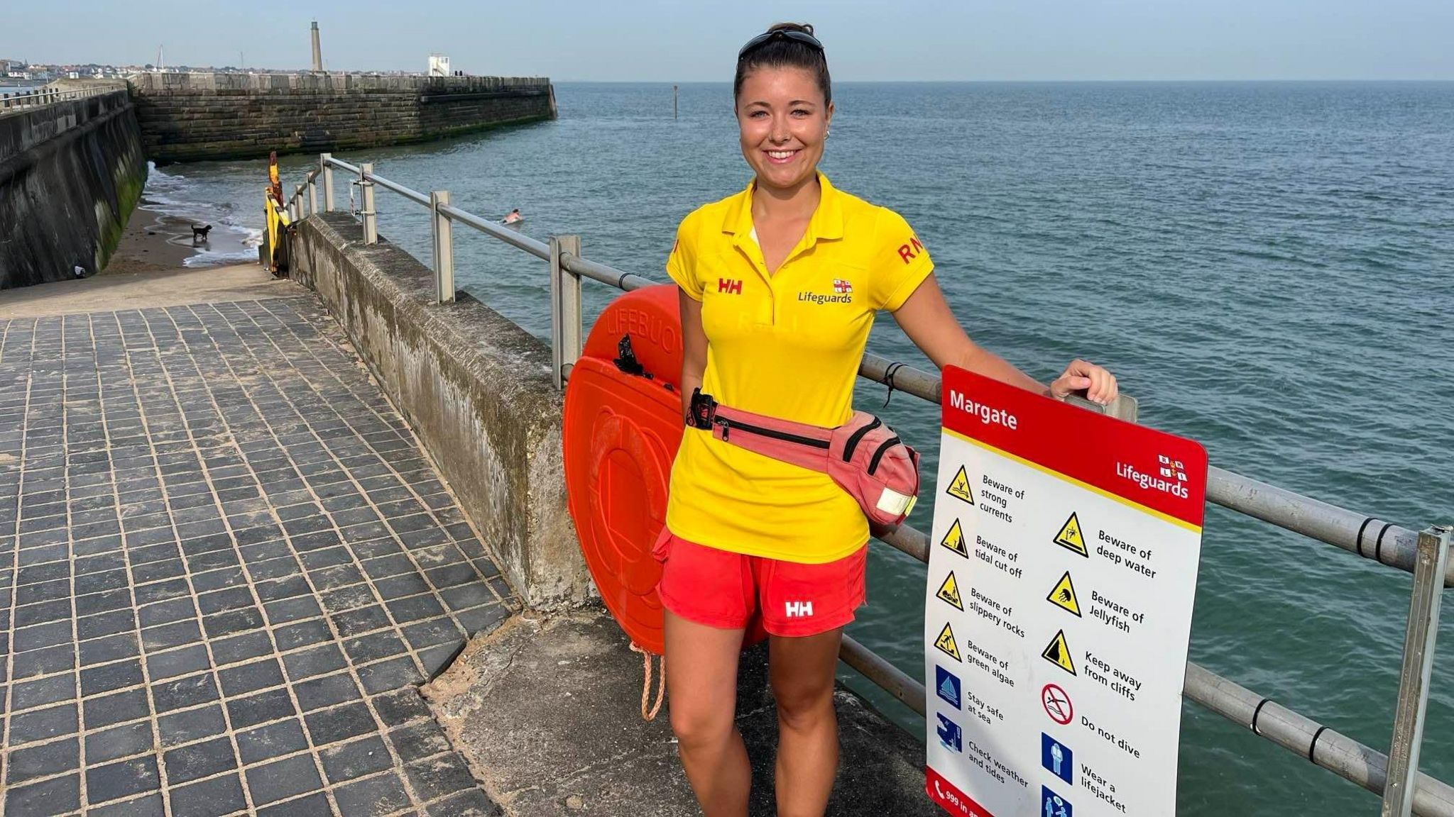 A RNLI lifeguard stands by metal railings with the sea behind her.