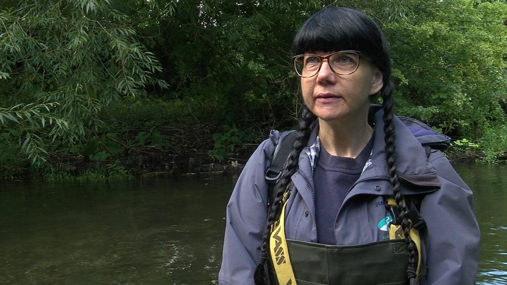 A woman with glasses and long black hair tied into two plaits, wearing a dark grey waterproof coat and black waders. There's trees and a river behind her.