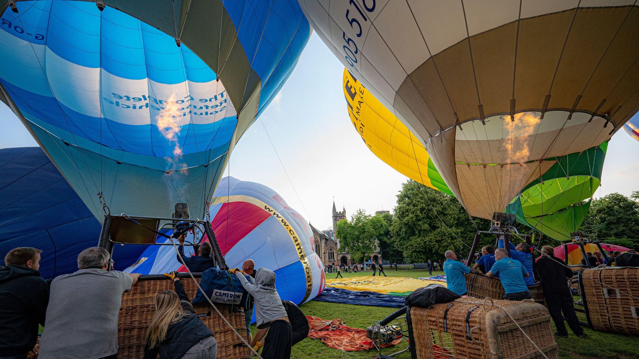 People inflating the hot air balloons using gases from the fire while others hold onto the baskets