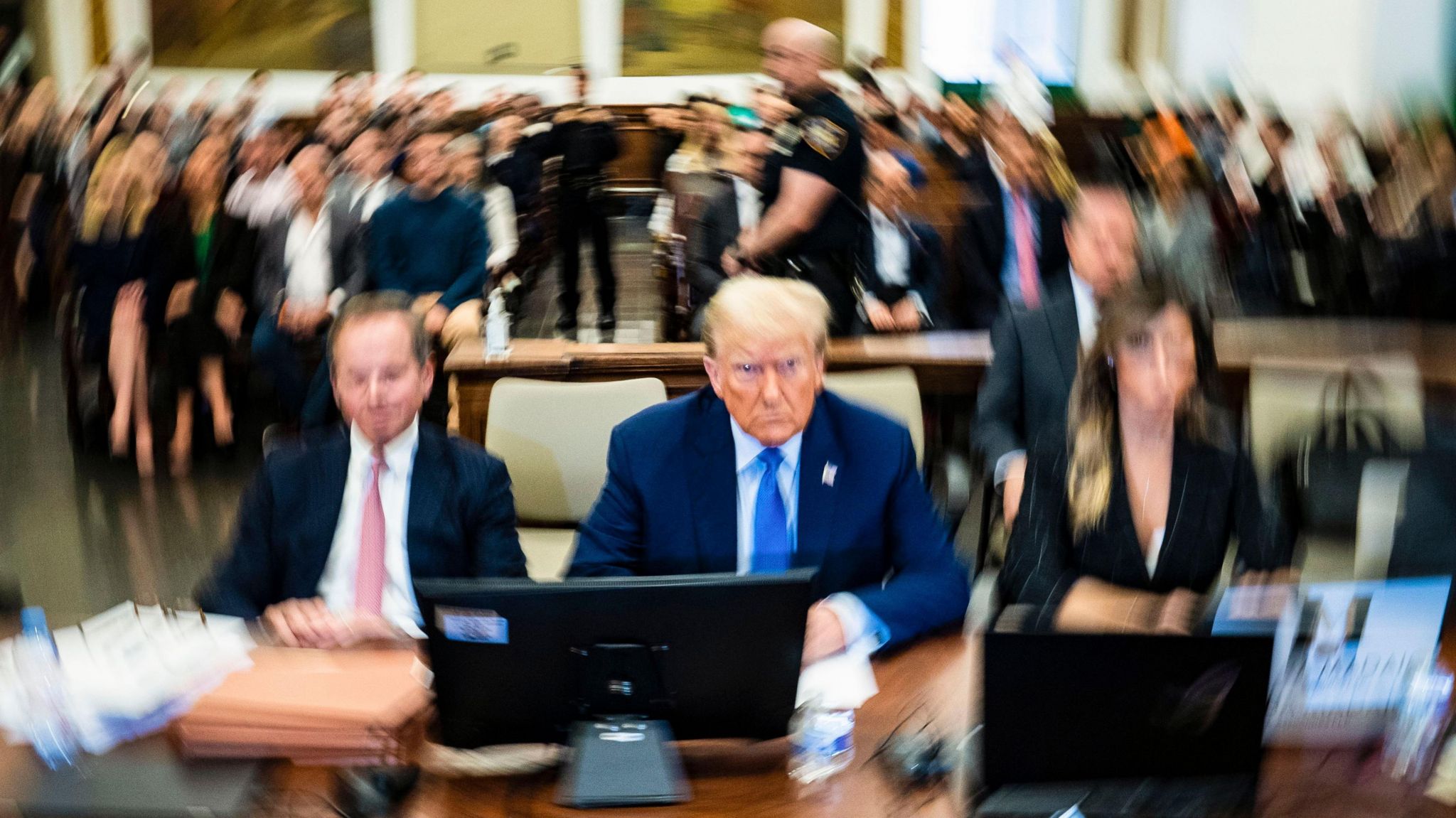 Donald Trump sits in court, flanked by his lawyers, with others in court behind him. Much of the image is blurry with Trump in focus in the centre.