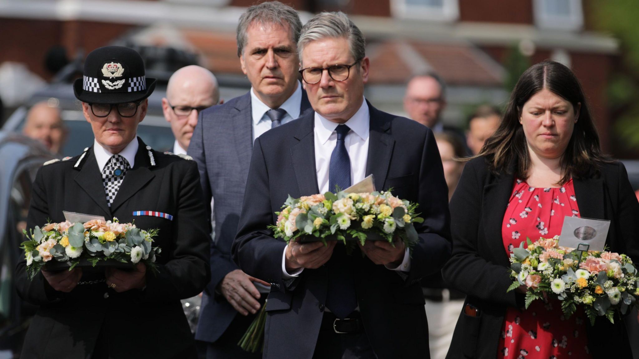 Merseyside Police Chief Constable Serena Kennedy, in full police uniform, Prime Minister Sir Keir Starmer, in a black suit, white shirt and black tie, and Merseyside Police and Crime Commissioner Emily Spurrell, in a red patterned dress and a black jacket, carry pink, peach and white floral wreaths