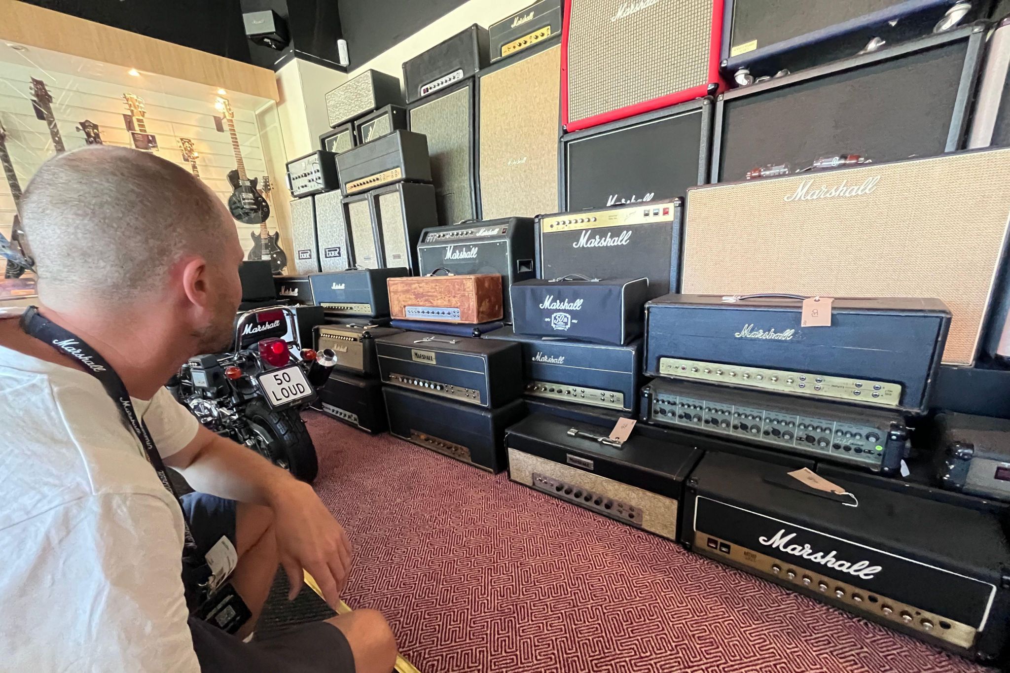 Rows of Marshall equipment lined up on a maroon patterned carpet, with guitars hung up on the far wall. A man in a white shirt with a black Marshall lanyard looks at the equipment