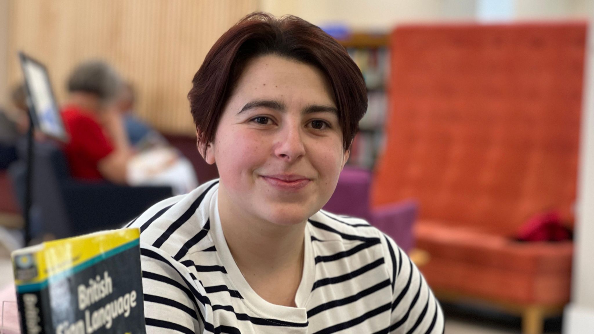 Ella smiles at the camera with a book on British Sign Language in front of her and behind her people are reading in the library