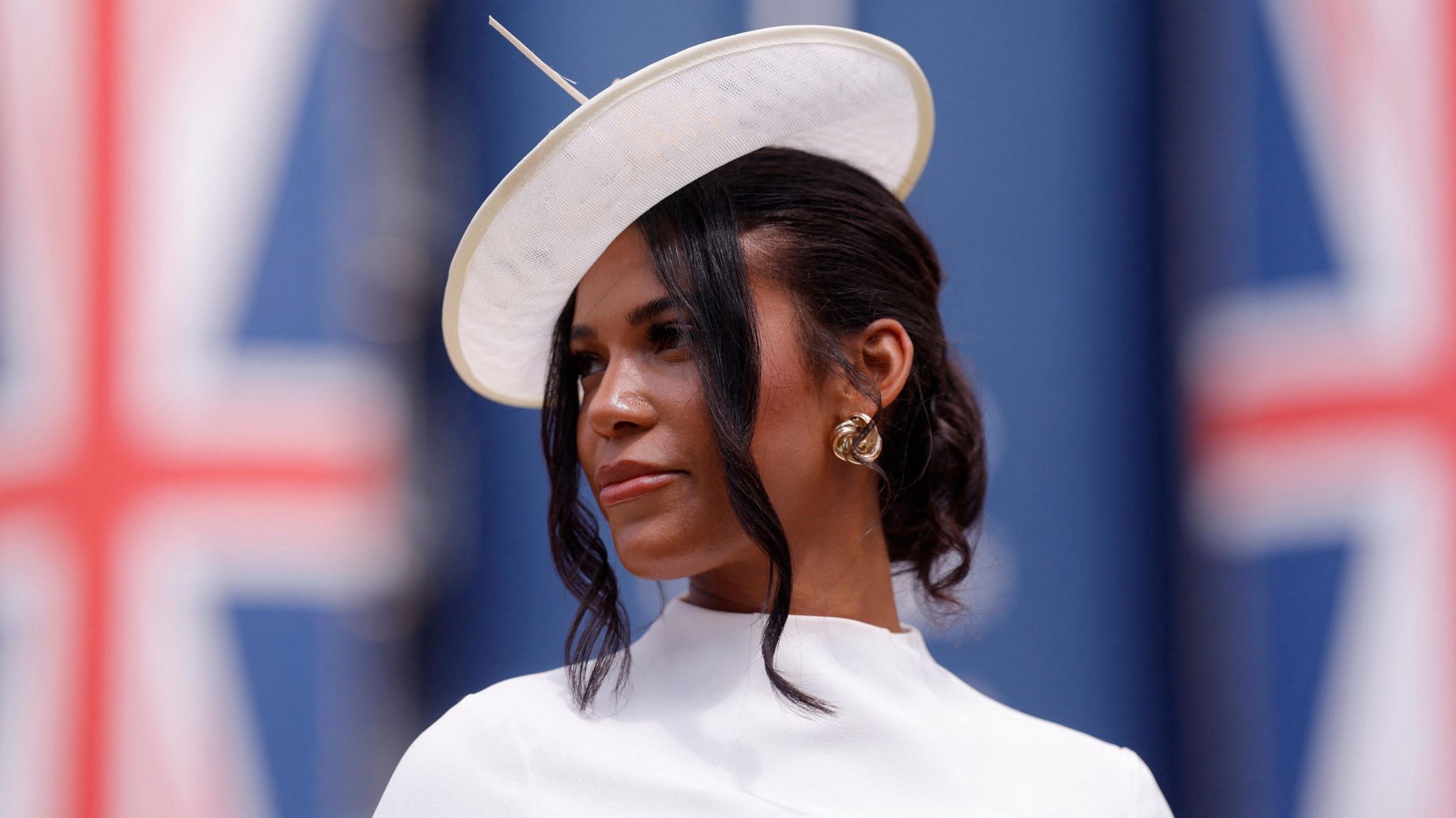 A woman looks away from the camera, wearing a wide brimmed white headdress, curled black hair in a messy bun and a high collar white blouse