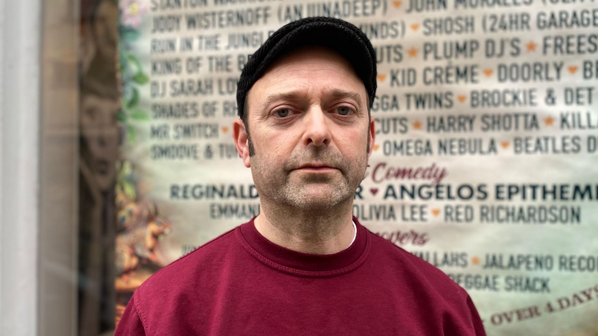 Simon Clarke wearing a red and gold Shindig Festival jumper standing in front of a big lineup poster