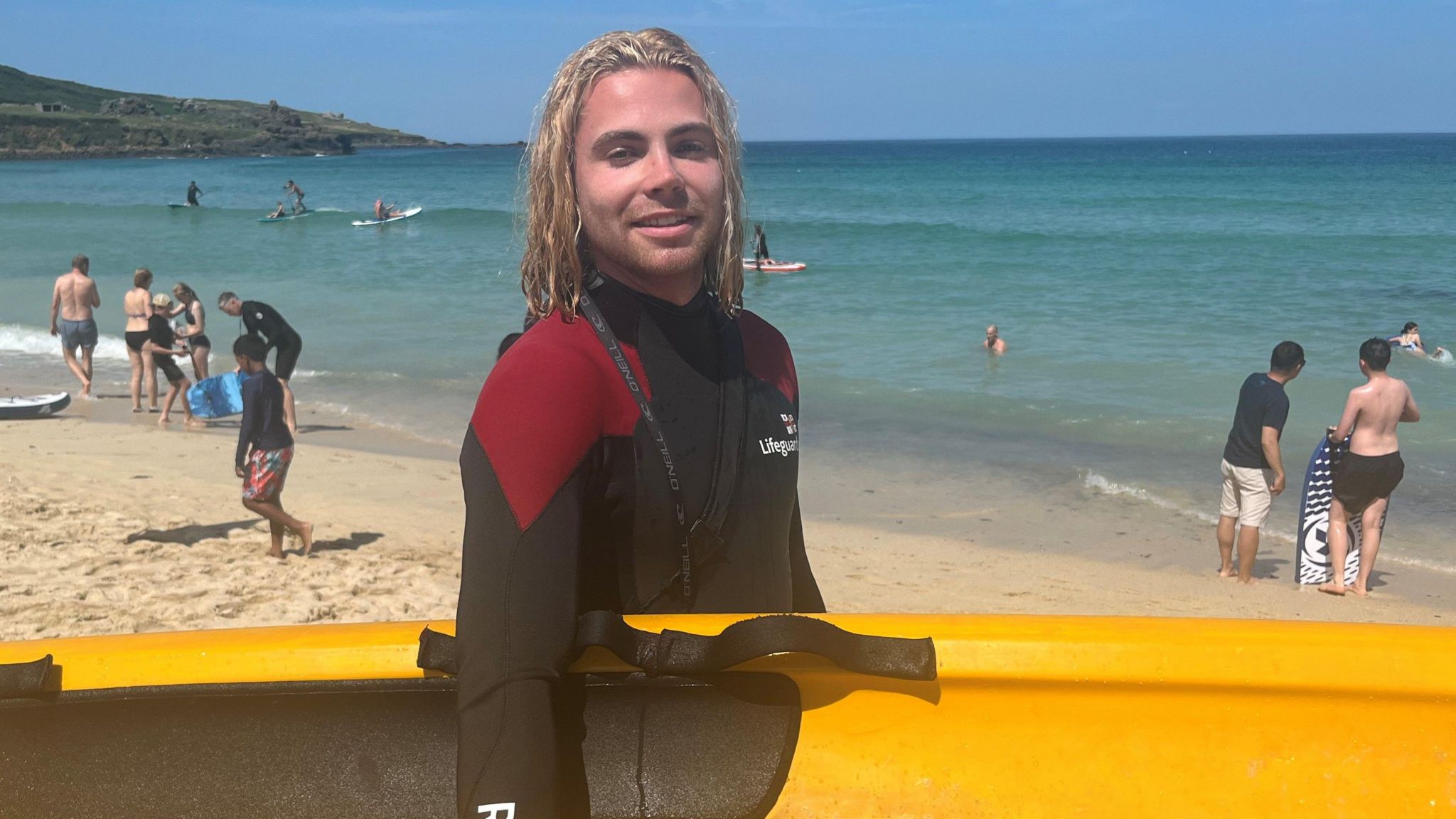 Lifeguard Reef Slack wearing a black and red RNLI-branded wetsuit carrying a yellow rescue board under one arm had shoulder length fair hair and is standing on the beach with the sea and swimmers, surfers and other beachgoers in the background