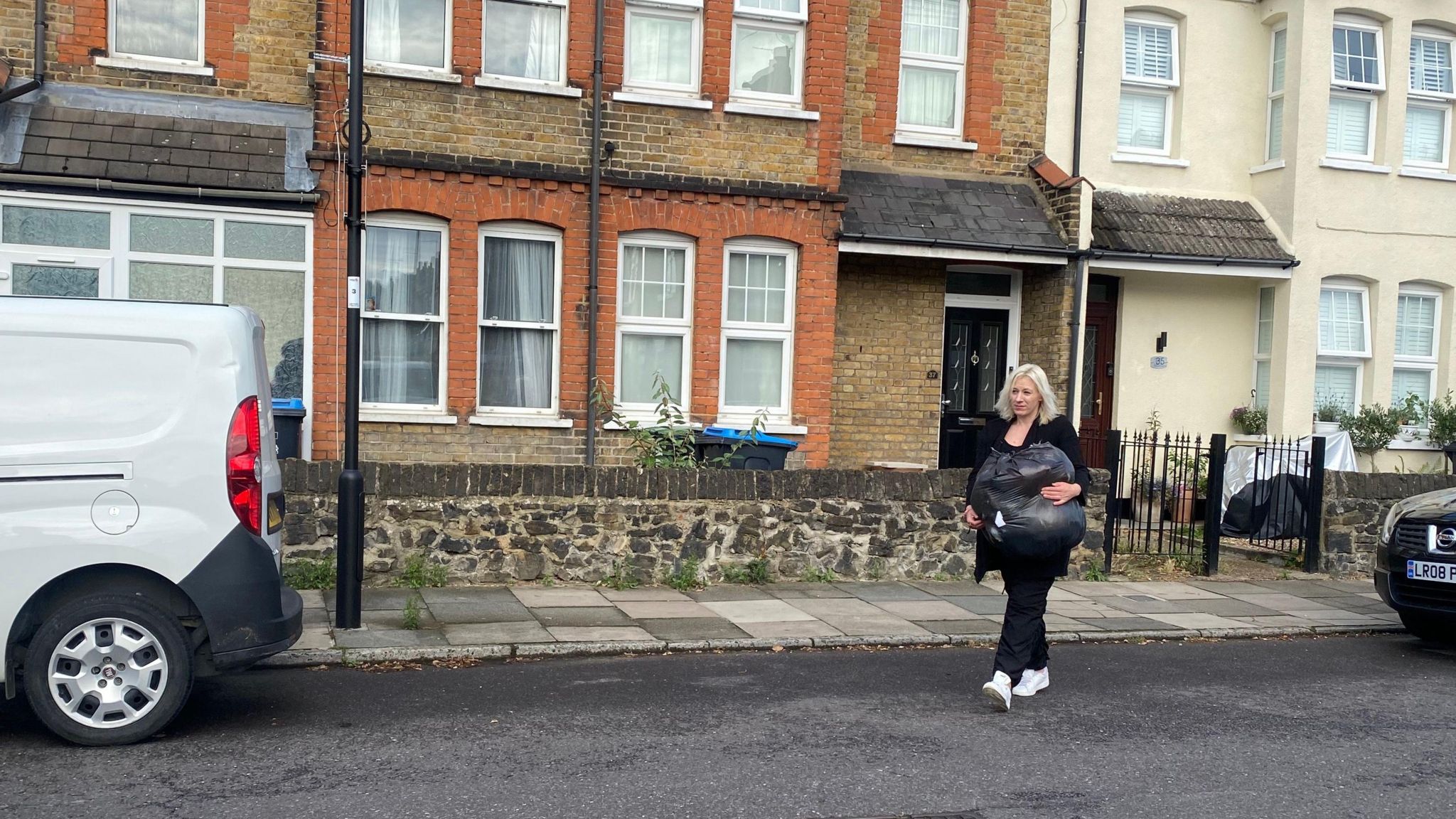 Carly, dressed in black top, trousers and jacket, walks crosses the road outside her former house while carrying a black bin bag