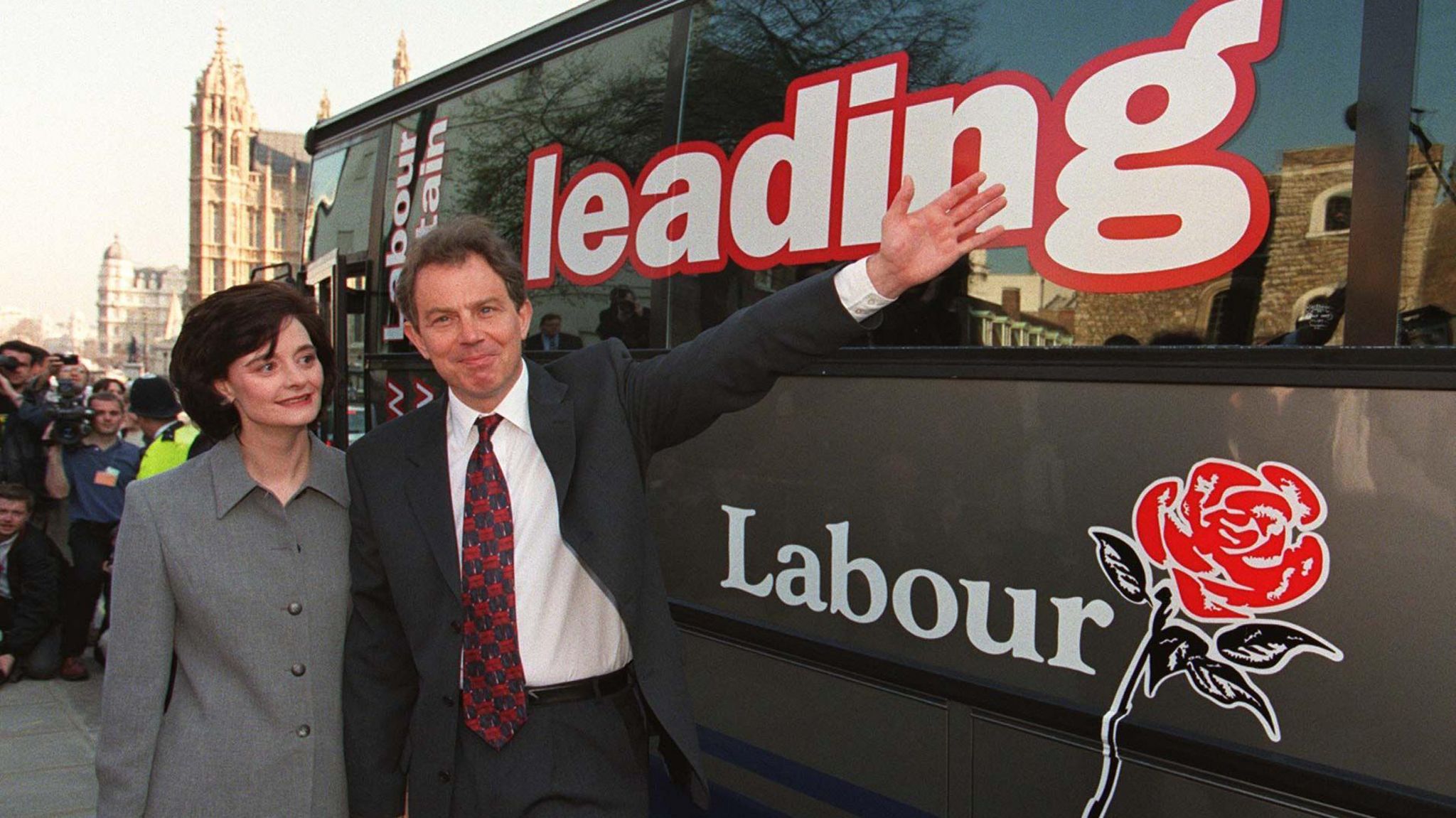 Tony Blair and his wife, Cherie, are pictured in Westminster stood in front of a large grey bus. Tony Blair is holding his arm up signalling towards the bus which is inscribed with 'leading Labour' on the side of it, along with a red rose that is the Labour Party's emblem.