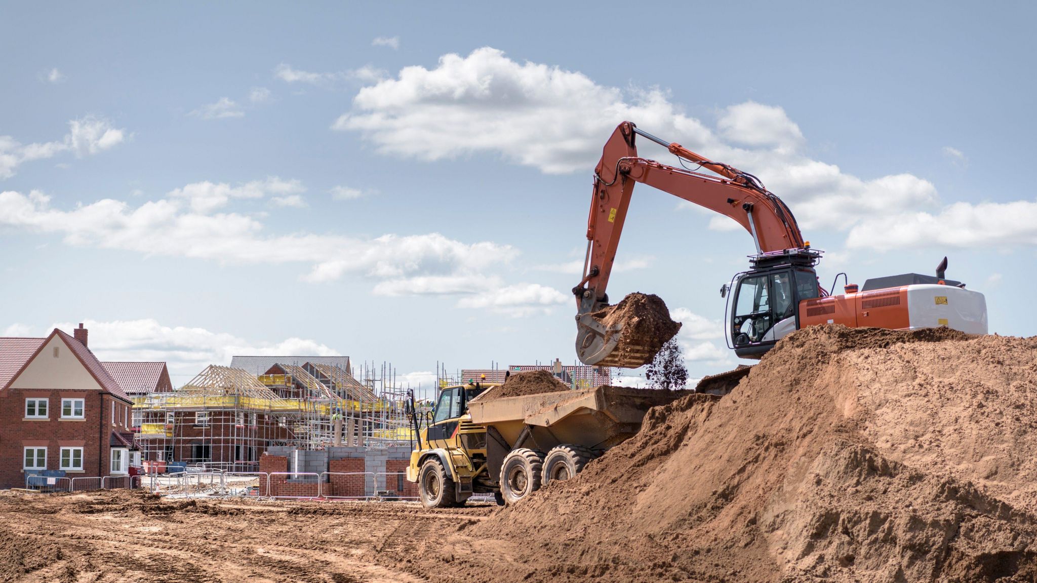 A stock image of a construction site. A digger can be seen on top of a large pile of earth, lifting it into the back of a truck. In the background there are new-build houses, some of them covered in scaffolding. 