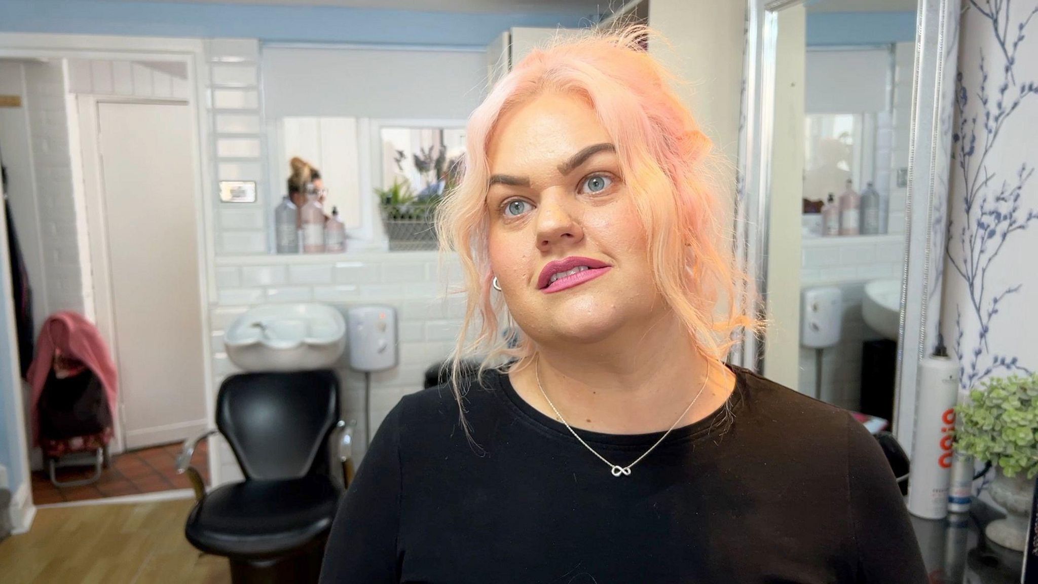 Kim Dudley stood in a hair salon with a chair and a basin behind her. She has blonde shoulder length hair with pink highlights and is wearing a black top with a thin silver necklace