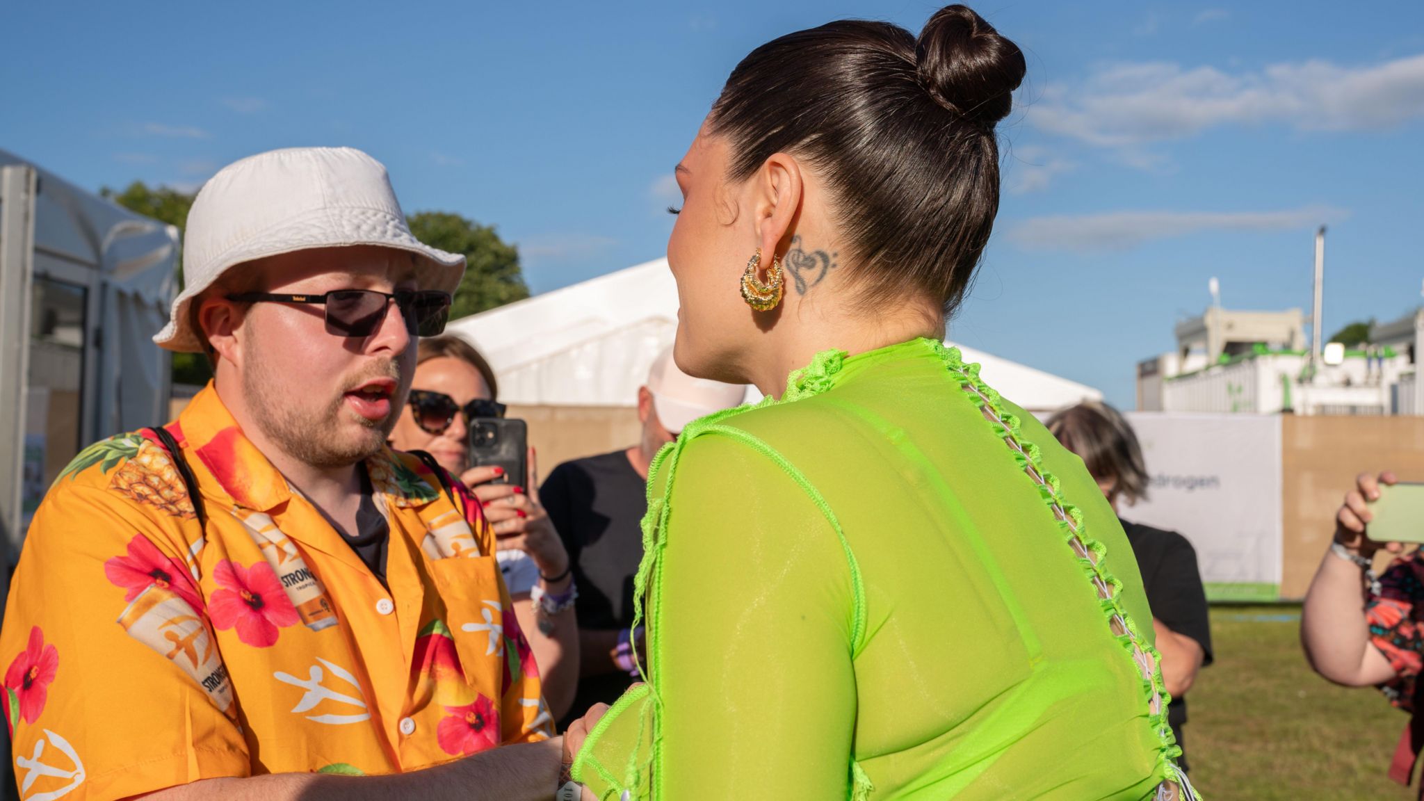 Jessie J wearing a green outfit staring and holding the hands of Nathan who is wearing an orange shirt with pink flowers printed on it, he is also wearing a white bucket hat and sunglasses on what is a sunny day backstage at the festival. People are surrounding the pair and are taking photographs of them using mobile phones.