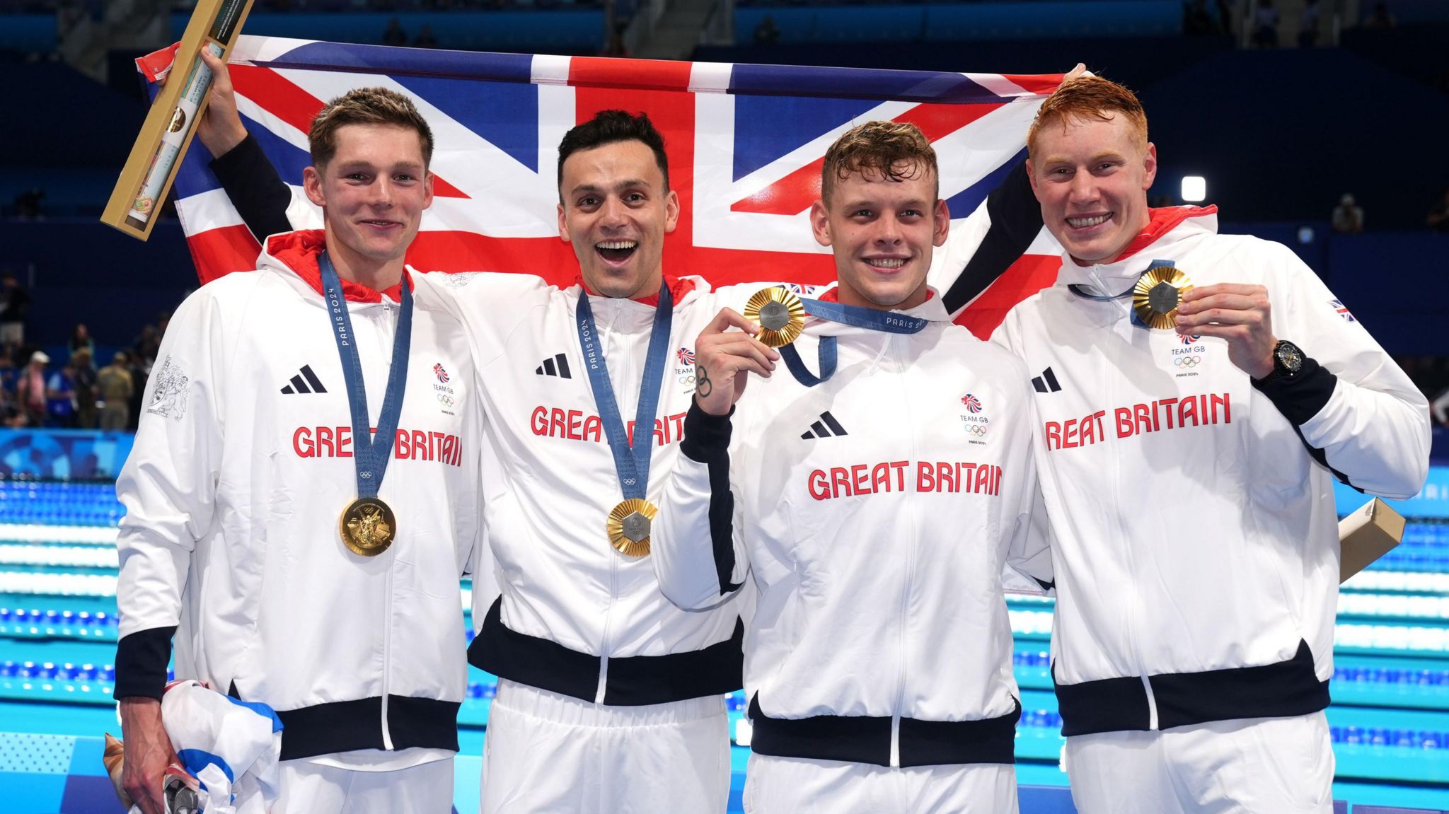 Duncan Scott, James Guy, Matthew Richards and Tom Dean. Duncan has short light hair, James has short dark hair, Matthew has short light hair and Tom has red short hair. They are all wearing white Adidas branded Great Britain jackets and wearing gold medals. They are holding a union jack behind them and smiling at the camera. 