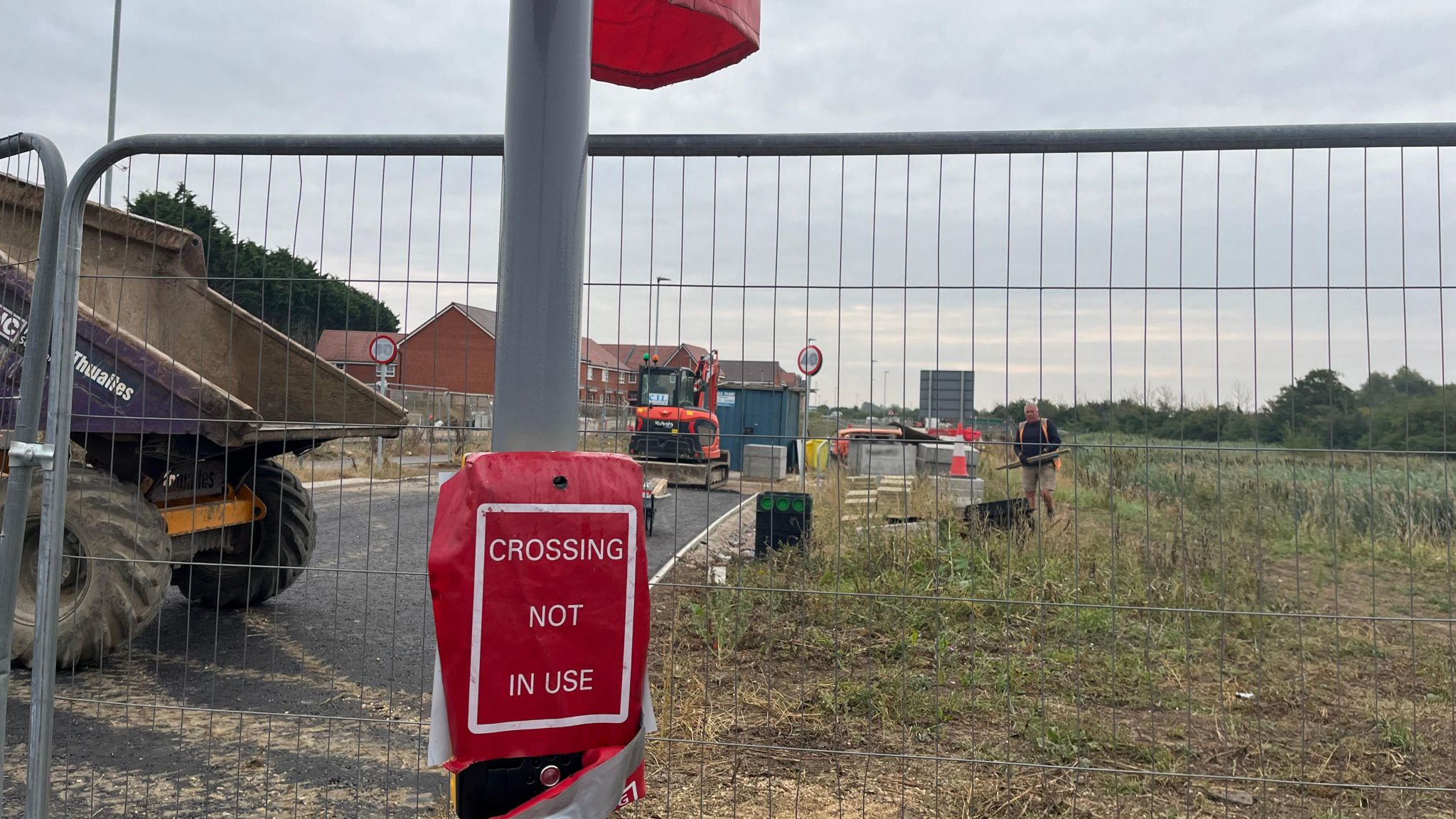 A post with a red box saying "crossing not in use", in front of a metal fence, grassland and highway equipment