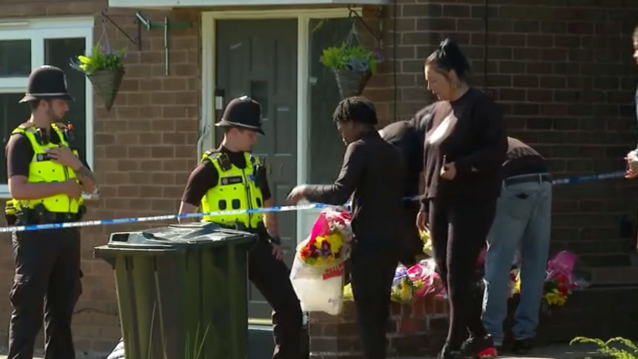 Two police officers standing outside a house. A man and woman wearing black are standing by a police cordon around the house holding flowers.