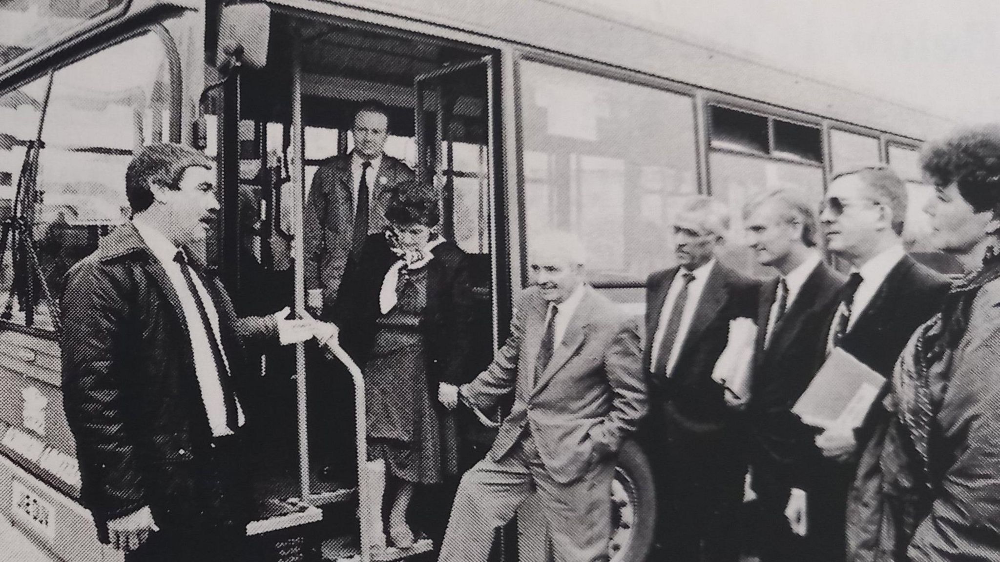 A black and white photo of a bus, with Kenny Beckers in a white shirt and black tie chatting to six men and a woman