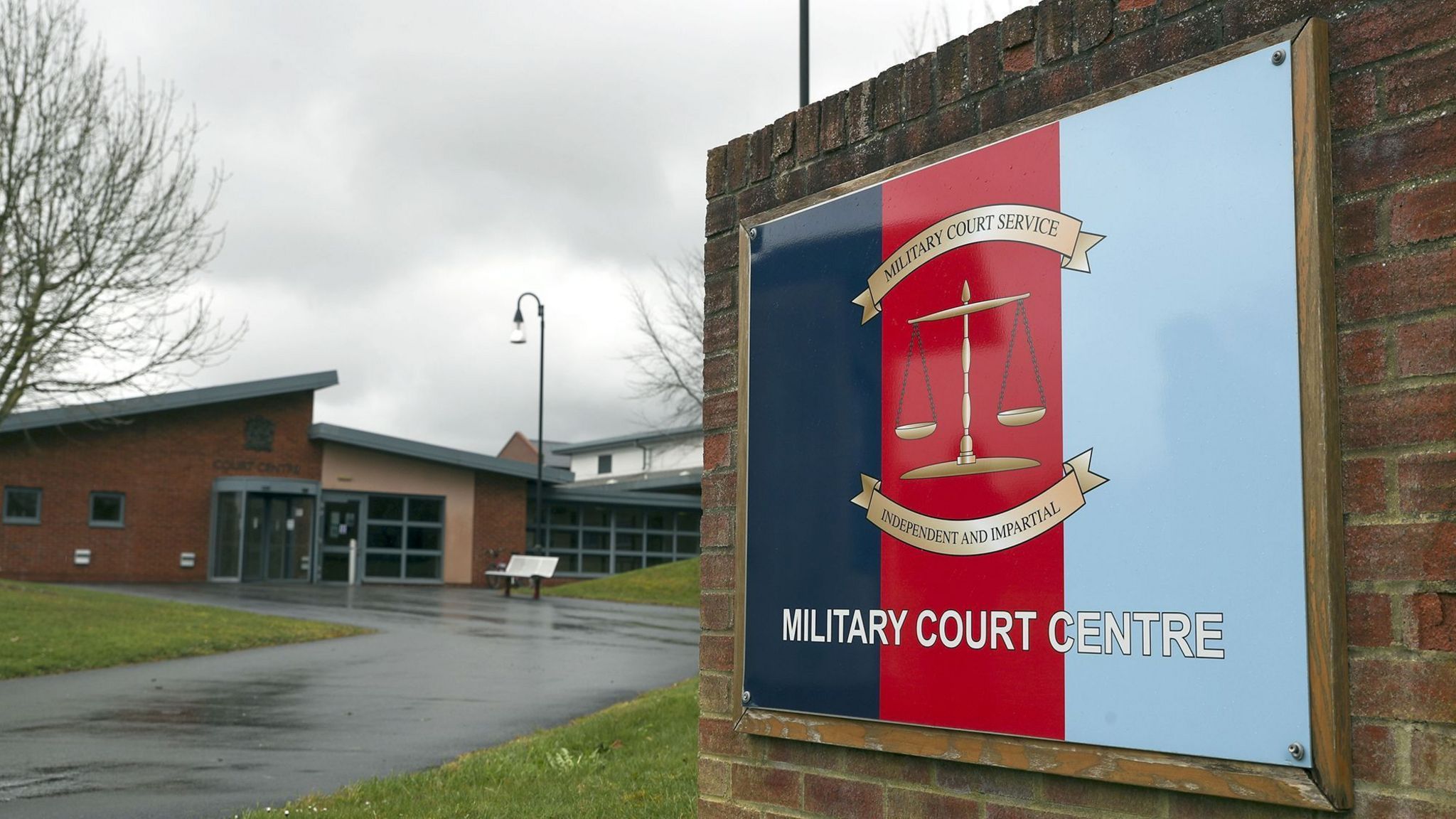 Entrance to Bulford Military Court Centre, with tricolour sign (navy blue, red and light blue to symbolise the navy, army and RAF). The sign reads 'Military Court Centre' and has a weighing scale in the middle, with scrolls above and below reading 'Military Court Service' and 'Independent and Impartial'