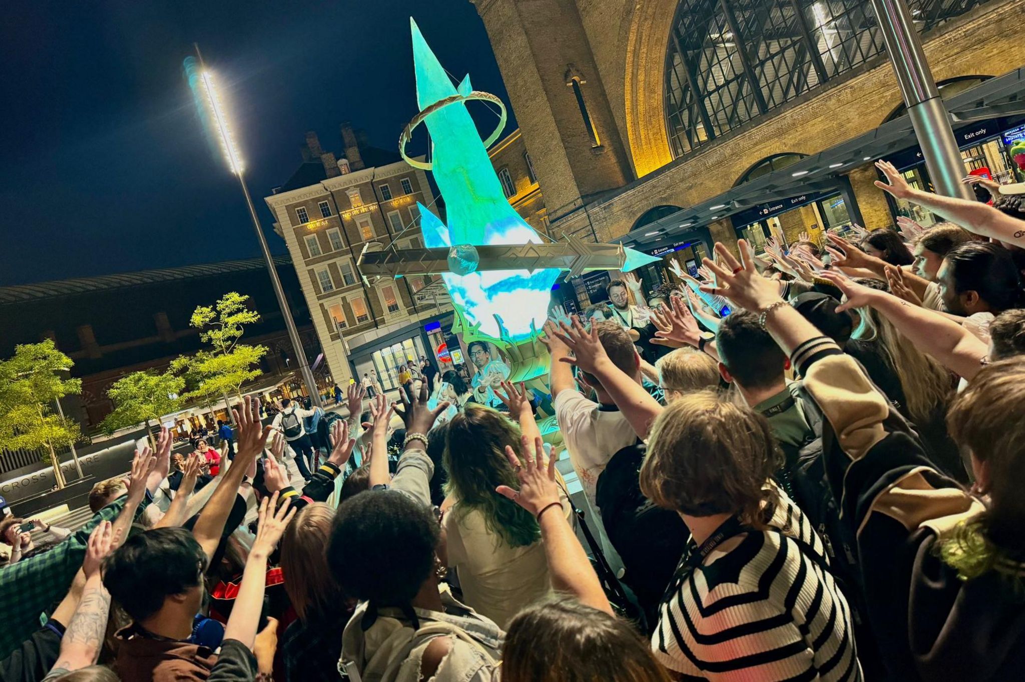 Dozens of people form a circle around a giant, glowing replica of a teal-coloured crystal. Everyone in the crowd has one arm outstretched towards the crystal, palm open and fingers splayed wide.