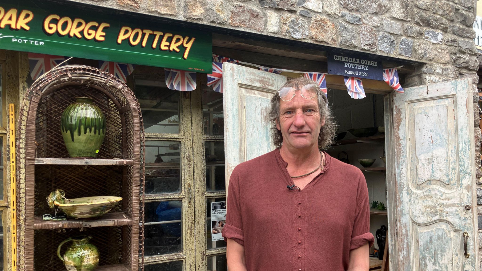 Man looking at camera in front of his pottery studio in Cheddar Gorge. He is wearing a red top and glasses are perched on top of his head