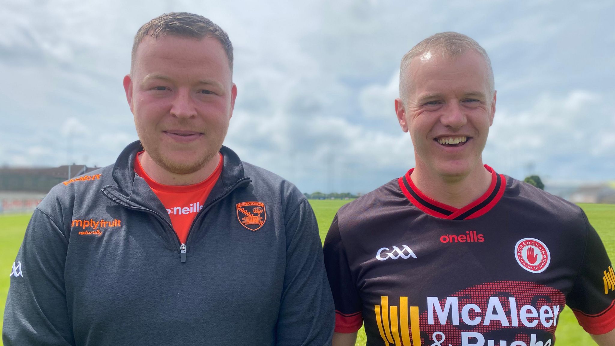 Two men smile directly into the camera. On the left Jamie is wearing a navy GAA branded quarter-zip top with a red jersey, Malachy is wearing a GAA jersey 