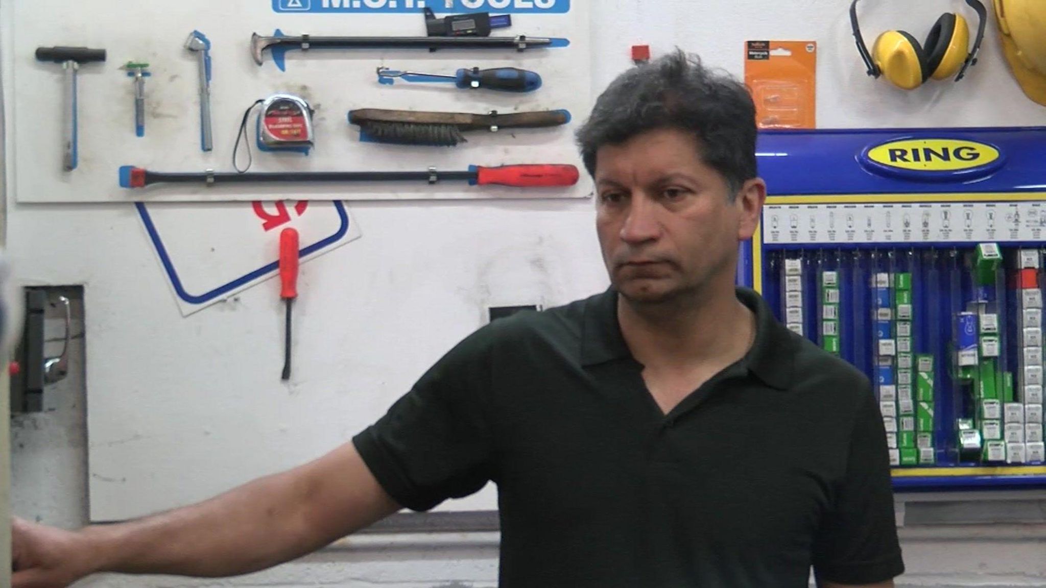 A man wearing a black t-shirt, with car garage equipment on the wall behind him