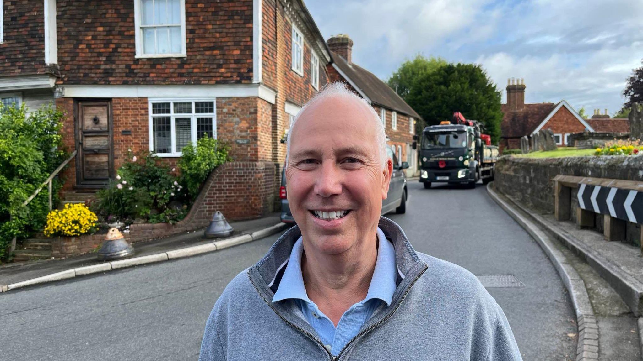 White haired man Kevin Rampling smiles at the camera while a small lorry squeezes round a small bend in Goudhurst