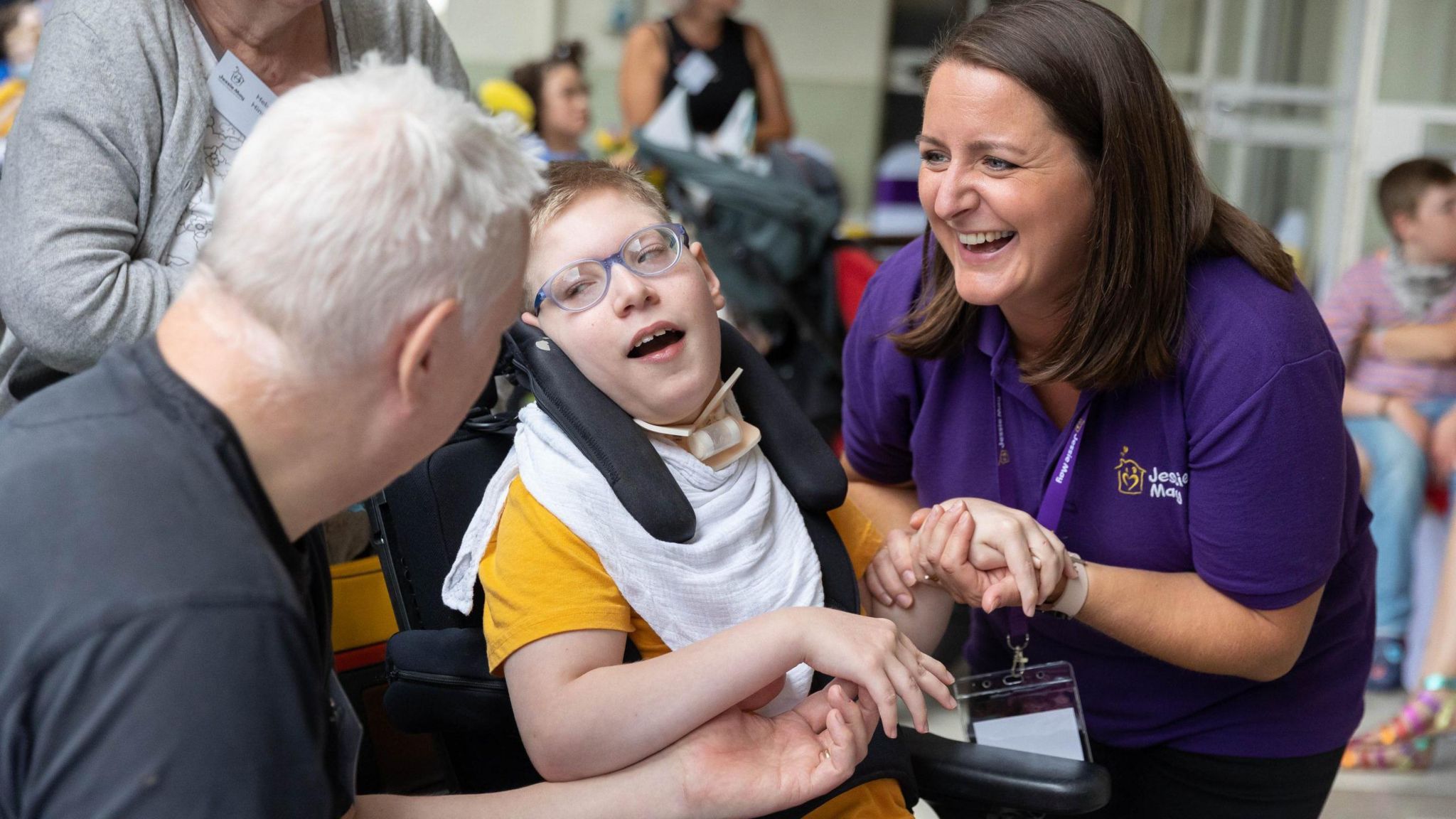 A young boy in a wheelchair holding hands with a nurse from Jessie May, who is smiling and wearing a purple t-shirt