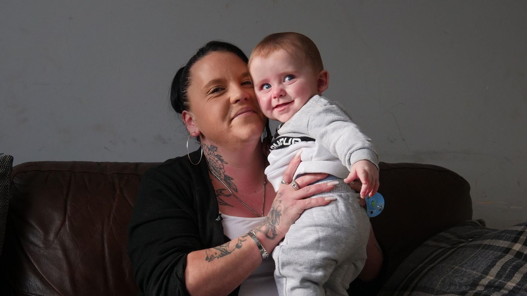 Kayleigh, a woman with dark hair and hoop earrings, sits on a sofa holding her baby. Kayleigh is looking into the camera and they are both smiling.