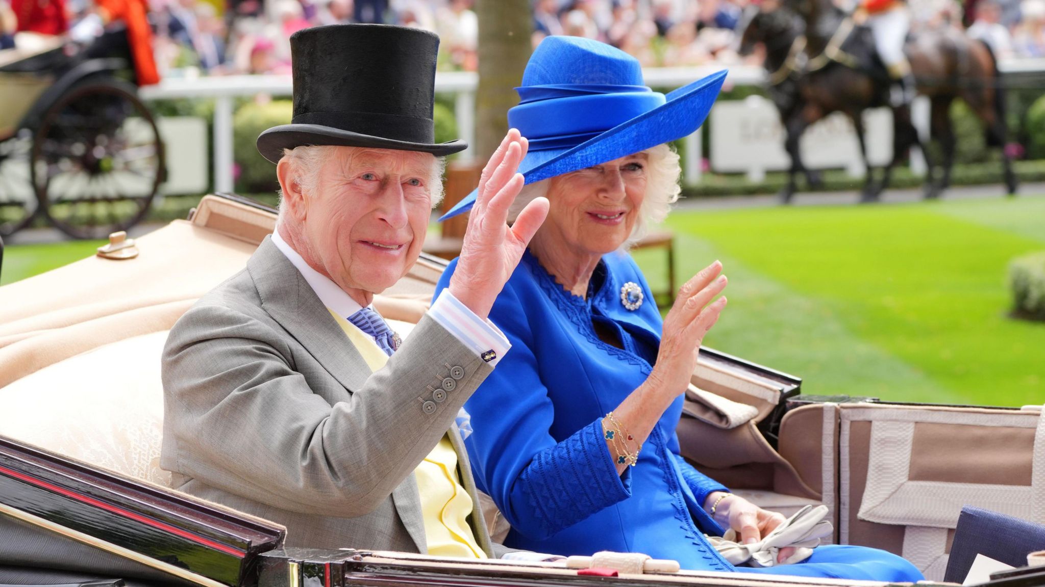 King Charles III and Queen Camilla waving at onlookers from a horse-drawn carriage