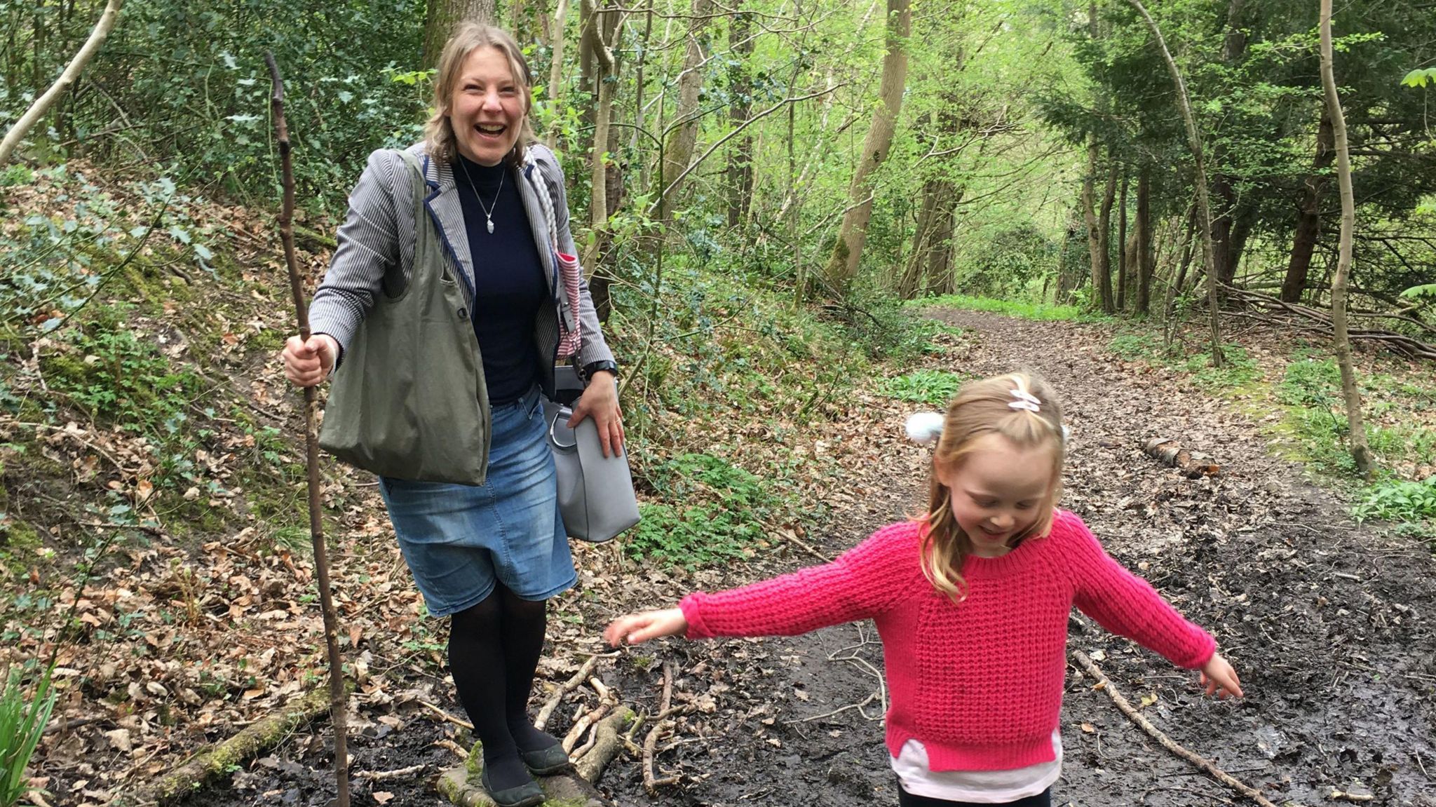 A smiling Mrs Clark, wearing a grey jacket, black top and blue denim skirt, holds a large stick as she walks in a woodland area with her young daughter who wears a red jumper.