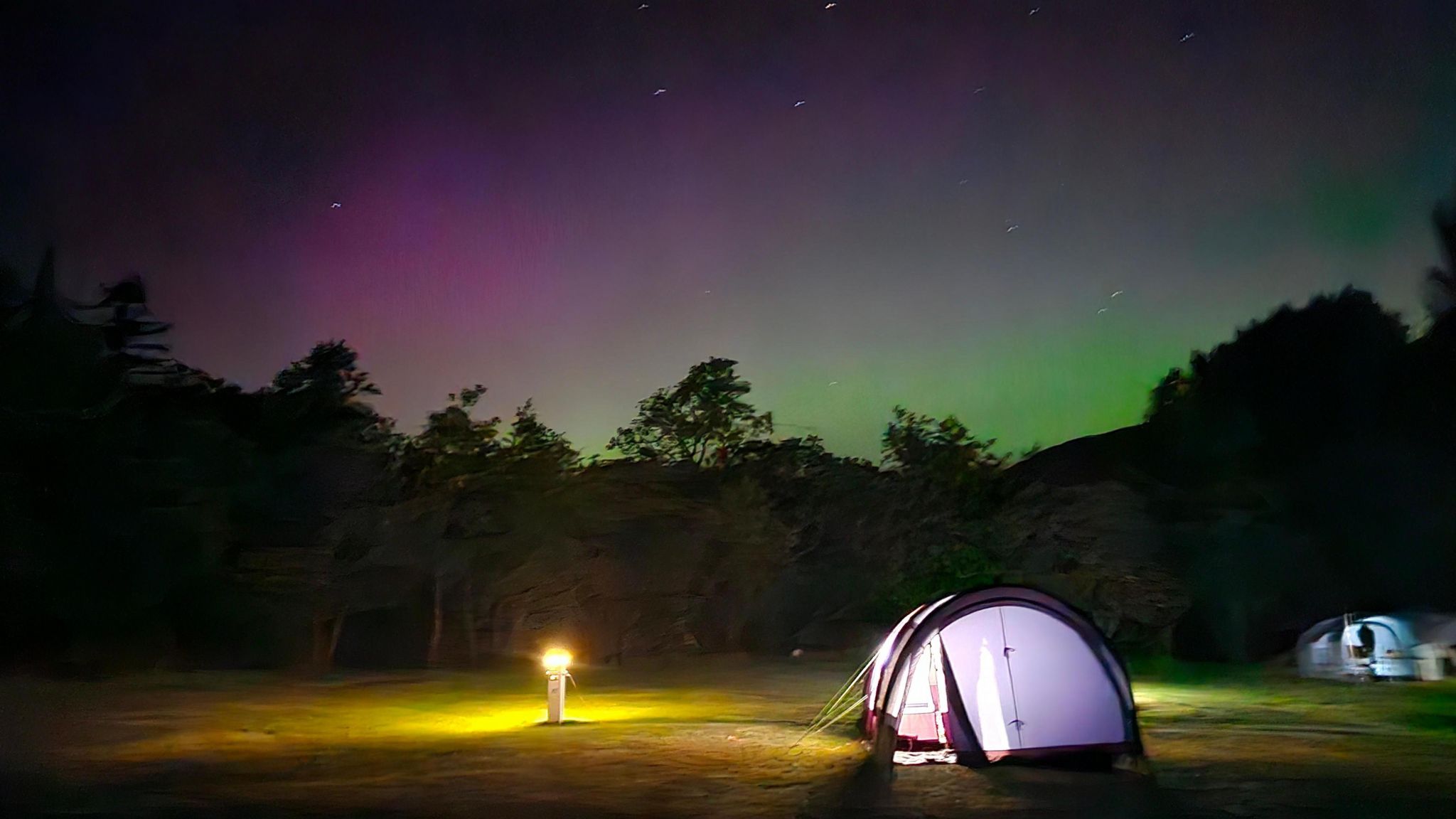 A tent in a small clearing in trees, with the sky glowing green and pink behind