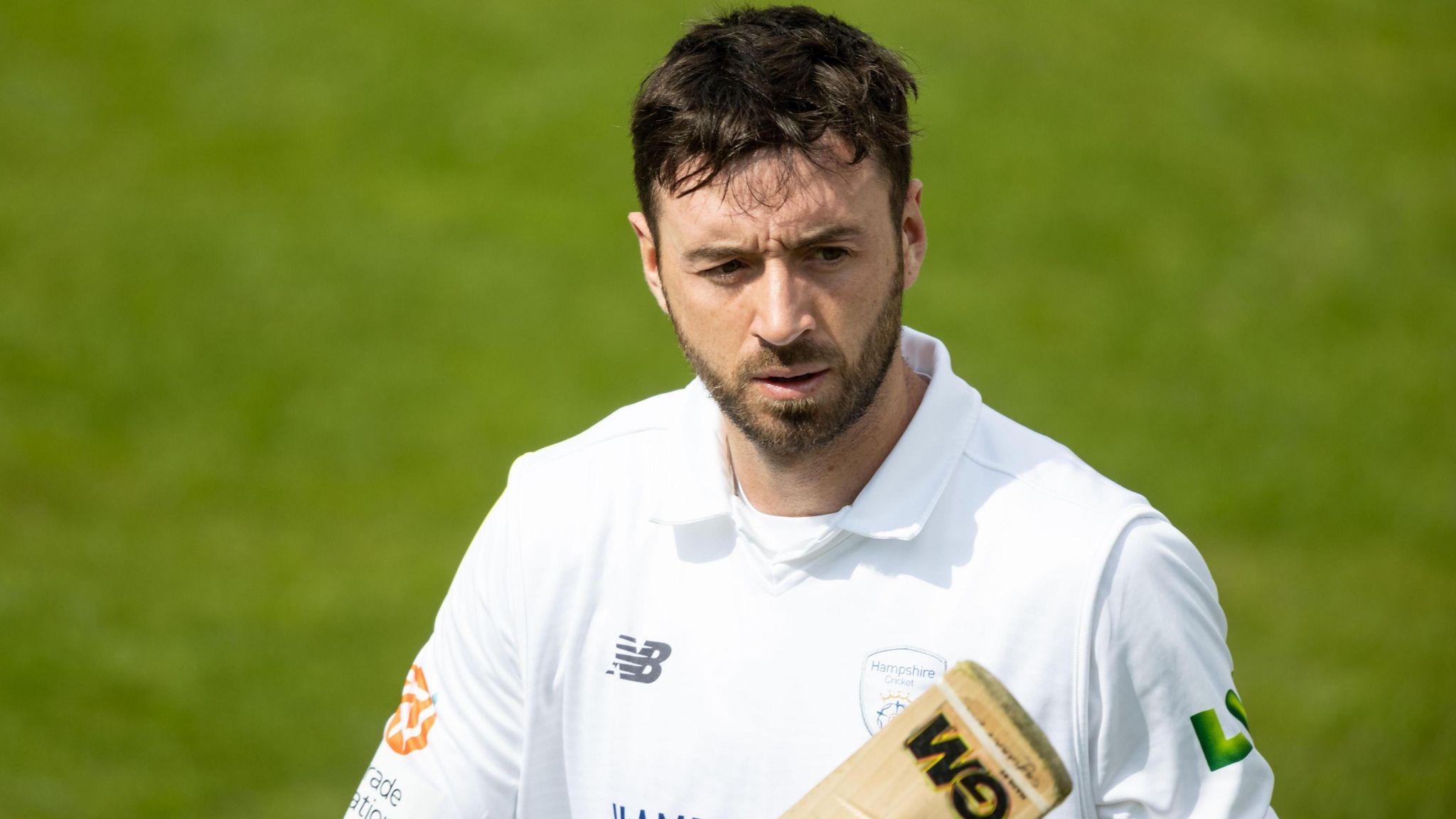 James Vince looks off camera while walking on the cricket pitch, he is wearing whites and carrying a cricket bat. He has dark brown short hair with a trimmed full beard.