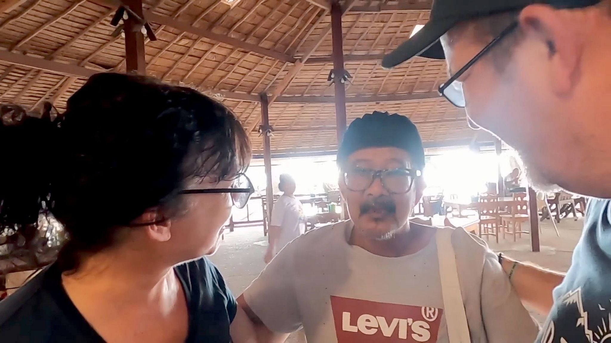 Woman and two men in a bar, the woman and one of the men are facing away from the camera and looking towards the other man, who is wearing a black hat and a grey t-shirt with a Levis logo in the centre. Behind them chairs and tables are visible